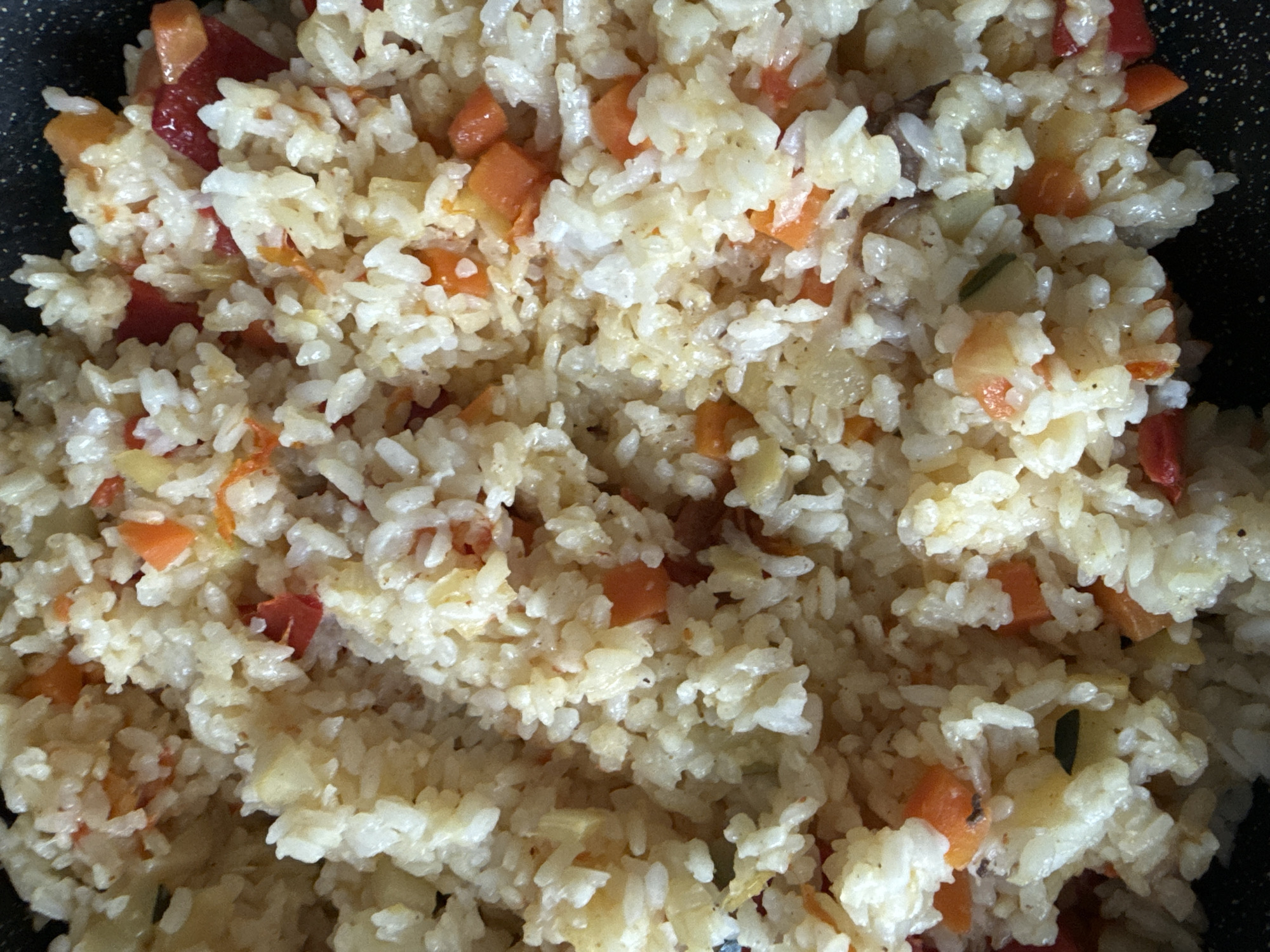 This media file is a close-up shot of cooked rice mixed with diced vegetables. The main subject is a bowl or pan of food, specifically rice pilaf or a similar dish. The rice grains are white and appear to be cooked until tender, with a slightly glossy sheen suggesting they've been prepared with oil or broth. Scattered throughout the rice are small, diced pieces of colorful vegetables. Predominantly visible are bright orange cubes, likely carrots, and some darker red pieces, possibly tomatoes or bell peppers. There are also lighter, yellowish-white pieces that could be onions or another root vegetable. A few dark green flecks, possibly herbs or small pieces of other vegetables, are also interspersed. The texture of the dish appears to be moist and well-mixed. The lighting is bright, suggesting it might be daytime, and the overhead perspective indicates the shot is taken from above the dish. There are no people, animals, or discernible background elements that provide specific location cues beyond the food itself. No text is visible in the image. The overall impression is of a home-cooked meal. Given the location context of Yakutsk, Russia, this dish could be a common meal or a special occasion dish prepared in that region. The food appears appetizing and ready to be served.