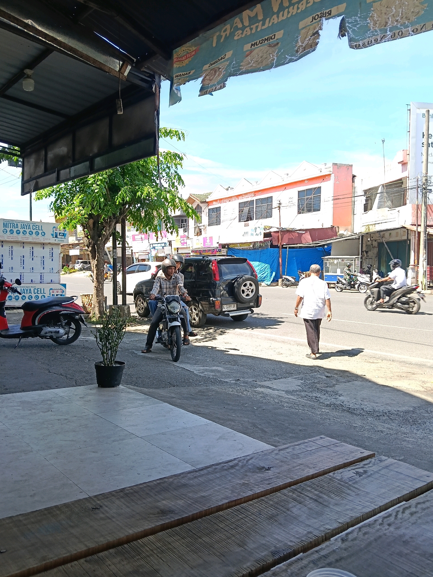 The image depicts a street scene in Banda Aceh, Indonesia, likely in a commercial area. In the foreground, a wooden table or bench is visible, with a tiled floor extending beyond it. To the left, a red scooter is parked. A black SUV is positioned in the middle of the street, with two young men on a motorbike beside it. One of the men is operating the motorbike, while the other is a passenger. Further down the street, a man in a white shirt and dark pants walks away from the camera, casting a shadow on the asphalt. Several buildings line the street, some appearing to be shops or businesses, with awnings and signage. A few other motorbikes are visible in the background, and the sky is clear blue with some scattered clouds. The overall impression is a busy, sunny day in an urban environment.