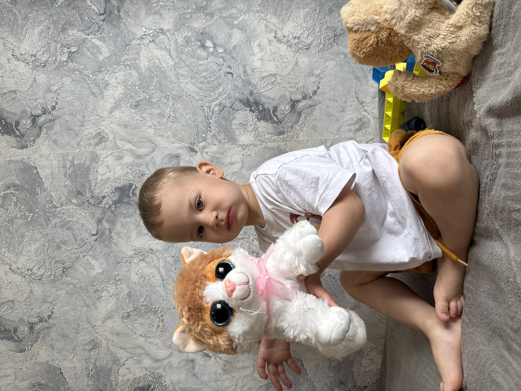 A toddler is sitting on a gray blanket, holding a stuffed cat toy with white and orange fur and a pink ribbon. The toddler is wearing a white t-shirt and yellow shorts. To the right of the toddler is a stuffed brown dog toy and a yellow and blue plastic toy. The background is a wall with a gray and white textured pattern. The image was taken indoors, in a location of Rusanovo, Russia.