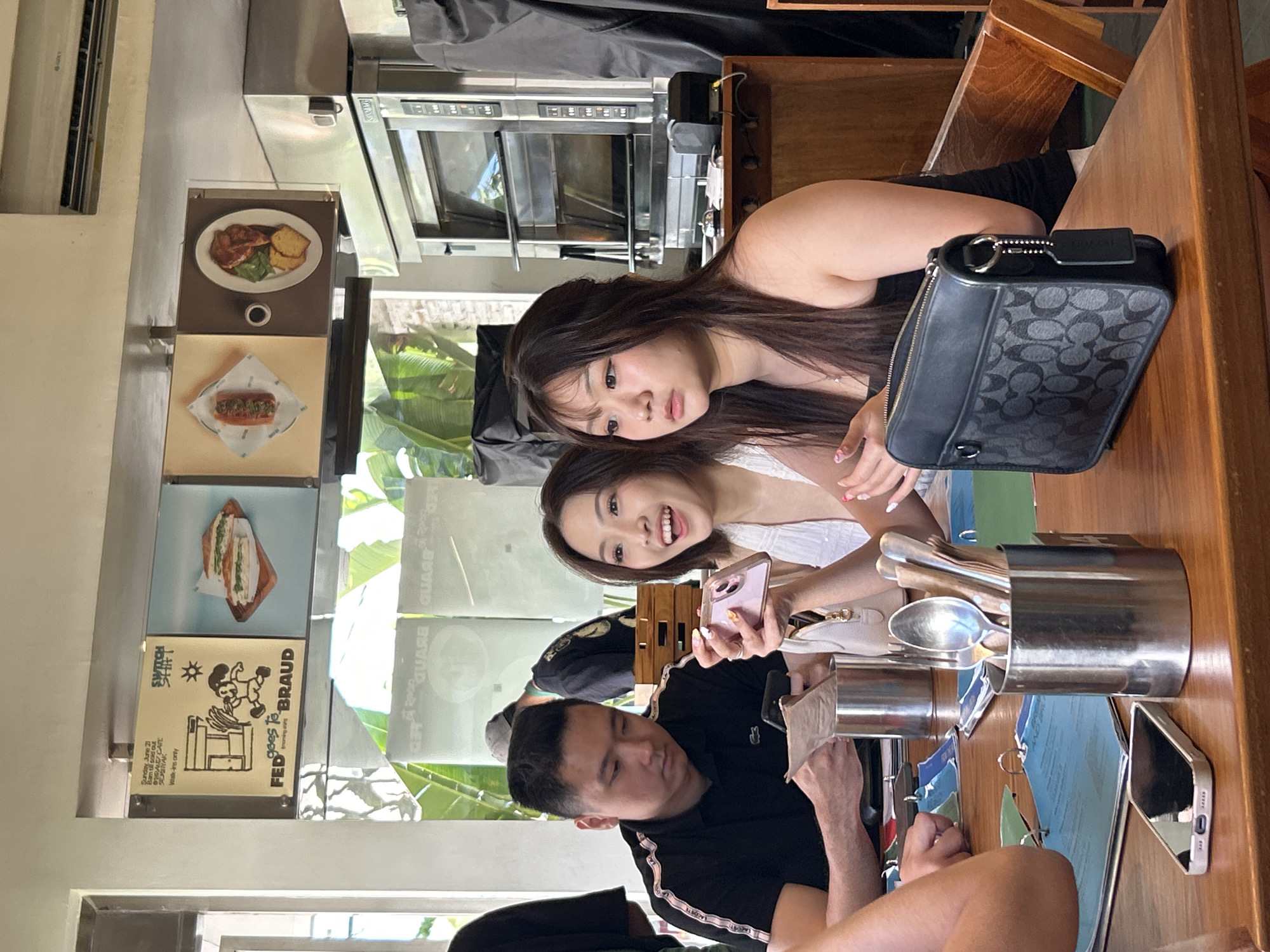 In North Kuta, Indonesia, at what appears to be a restaurant, three individuals are seated at a wooden table. A man in a black polo shirt is looking down at a paper. To his right, a woman with an excited expression is holding up a pink smartphone. Next to her, a woman with long brown hair is looking away from the camera with a neutral expression. A black Coach handbag is on the table in front of her. The background includes signage with images of food and a commercial oven. The environment suggests a casual dining setting, possibly a cafe or restaurant, with a warm, indoor ambiance.
