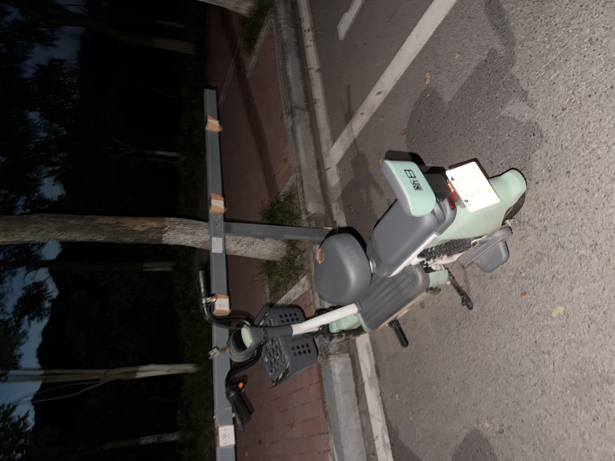 This image depicts a parked electric scooter in an outdoor setting at dusk or night. The scooter, light green and gray, has a basket on the handlebars and a license plate on the rear. It is positioned on an asphalt surface next to a red brick pathway and a concrete curb. Behind the scooter, a metal barrier with orange reflectors is visible, and several trees with visible bark stand in the background. The sky is dark, with hints of blue, suggesting twilight. The environment appears to be a public area, possibly a park or a street. The city of Jiangjin, China, is mentioned as the location, suggesting this scene is within its jurisdiction. There are no people or animals visible in the image, and no actions are taking place.