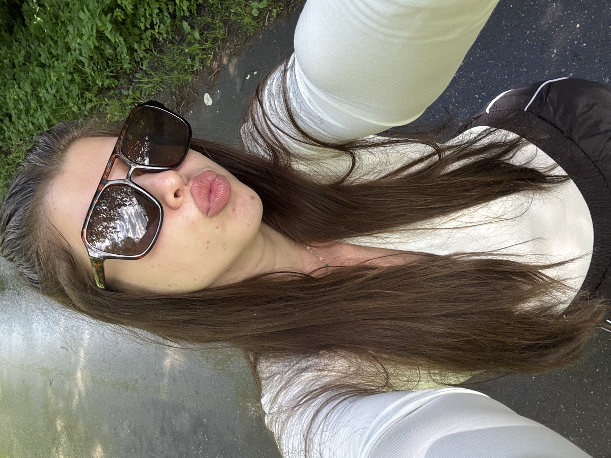 A selfie taken outdoors in daylight shows a young woman with long brown hair. She is wearing sunglasses and puckering her lips. She is wearing a long-sleeved white shirt and appears to be near a paved area with greenery in the background. The location in the city of Шевлягино, Russia, is not discernable from the image.