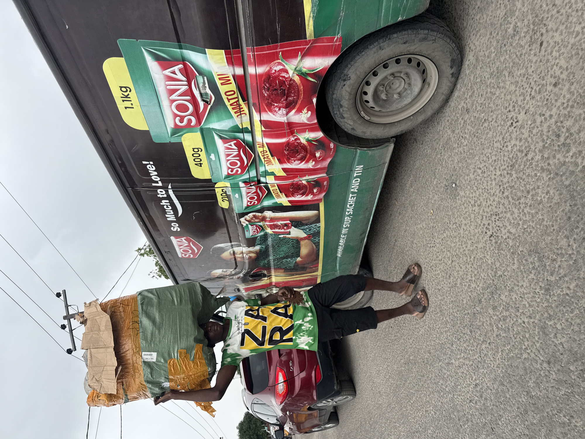 In Ado, Nigeria, a young man carries a large bundle of goods on his head while standing next to a dark-colored van. The van is decorated with a large advertisement for "SONIA" tomato paste, featuring images of tomato paste packets and ripe tomatoes, along with the slogan "So Much to Love!". The advertisement also lists various package sizes, including 1.1kg, 400g, and 210g, and states "AVAILABLE IN CUP, SACHET AND TIN". The man is wearing a green, white, and yellow t-shirt with the word "ZARA" printed on it, black cargo shorts, and brown sandals. He is holding a small object in his right hand. To the left of the man is a red SUV. The scene appears to be on a paved street with visible power lines overhead. The sky is overcast.