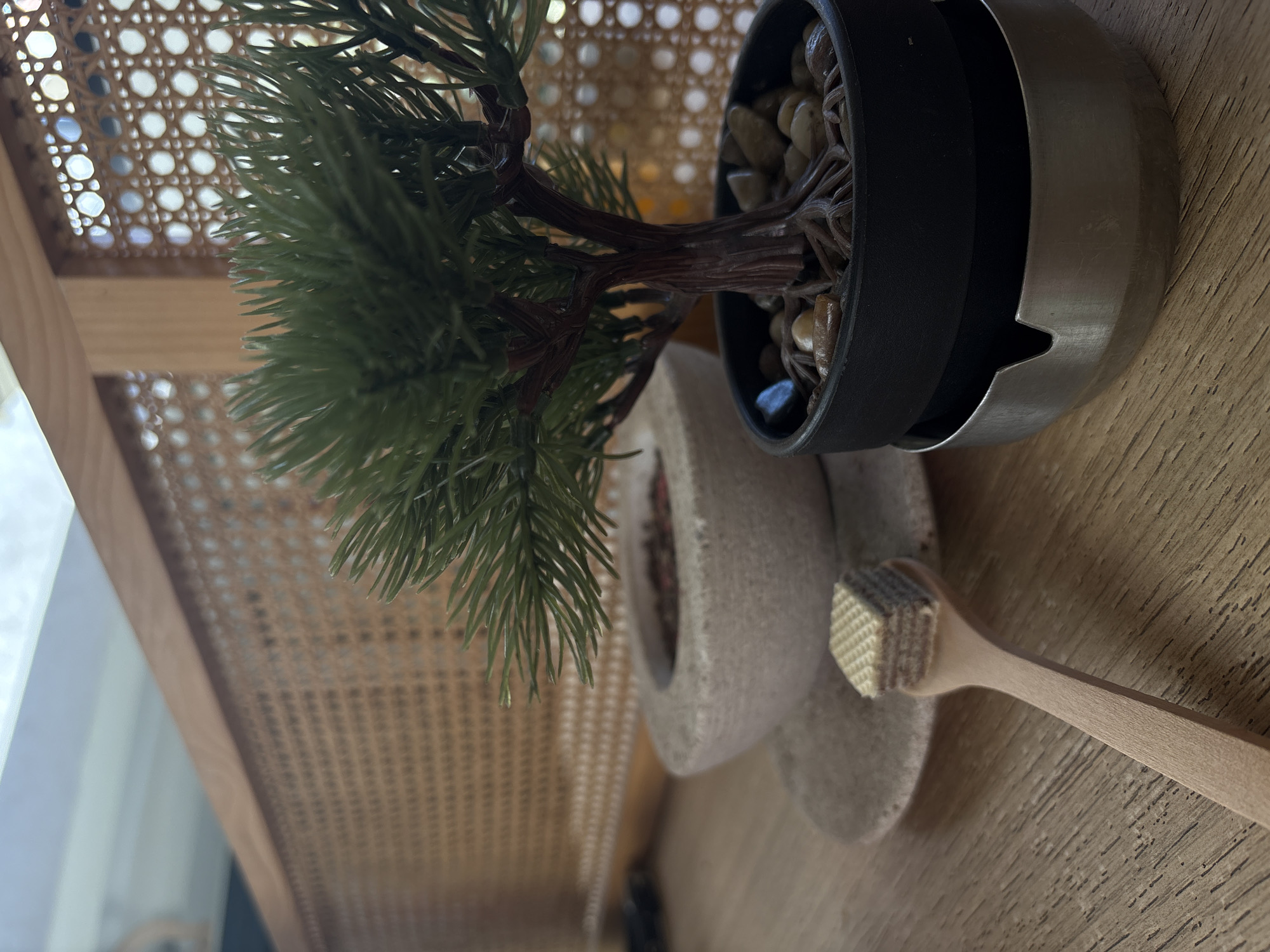 The image shows a small, artificial bonsai tree in a black and silver pot placed on a beige, flower-shaped coaster. To the right of the coaster is a wooden honey dipper. The bonsai tree has green needles and a brown trunk. The pot contains small pebbles. The coaster and dipper are resting on a wooden surface. In the background, there is a woven cane texture that could be a chair back or panel. The location is not discernible from the image. There is no information available to indicate that this image was taken in the city of Kragujevac, Serbia.