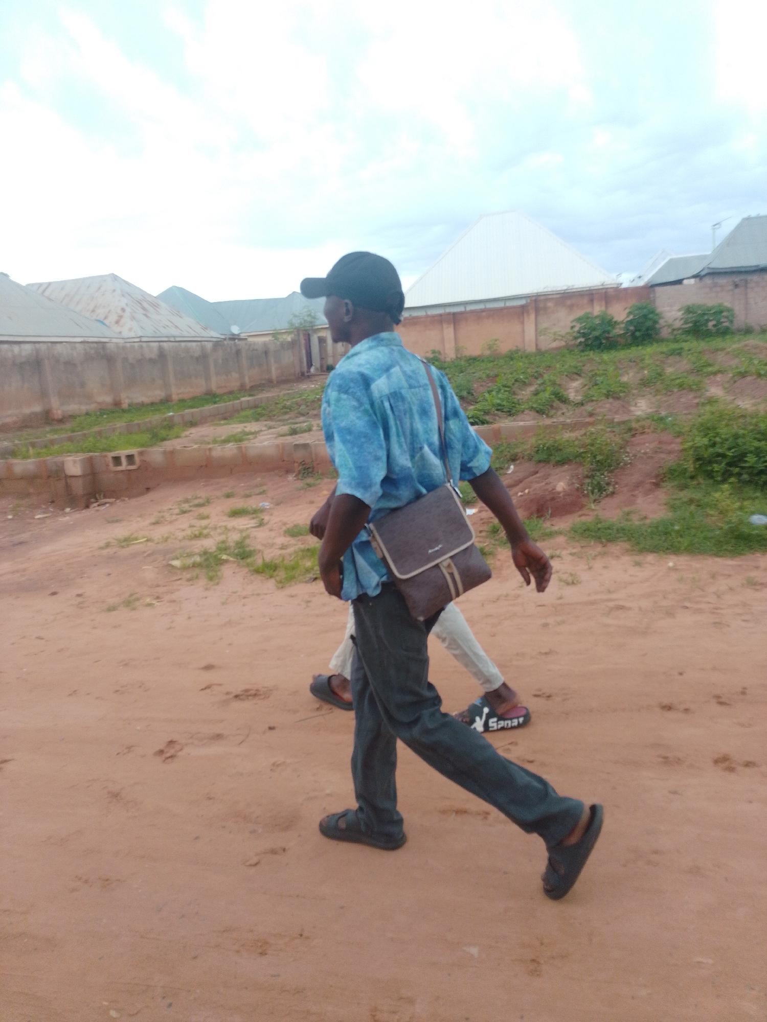 The image shows two individuals walking on a dirt road in the city of Tempure, Nigeria. The person in the foreground, wearing a blue patterned shirt and dark pants, is carrying a brown messenger bag. They are walking towards the right of the frame, with their body angled away from the camera. A second individual, partially visible behind the first, is also walking in the same direction. The background consists of a low concrete wall, behind which are several buildings with corrugated metal roofs, indicative of a residential area. Green vegetation is present along the roadside and behind the wall. The ground is a dusty, reddish-brown color, with some scattered debris. The sky is overcast with a cloudy, pale blue hue.