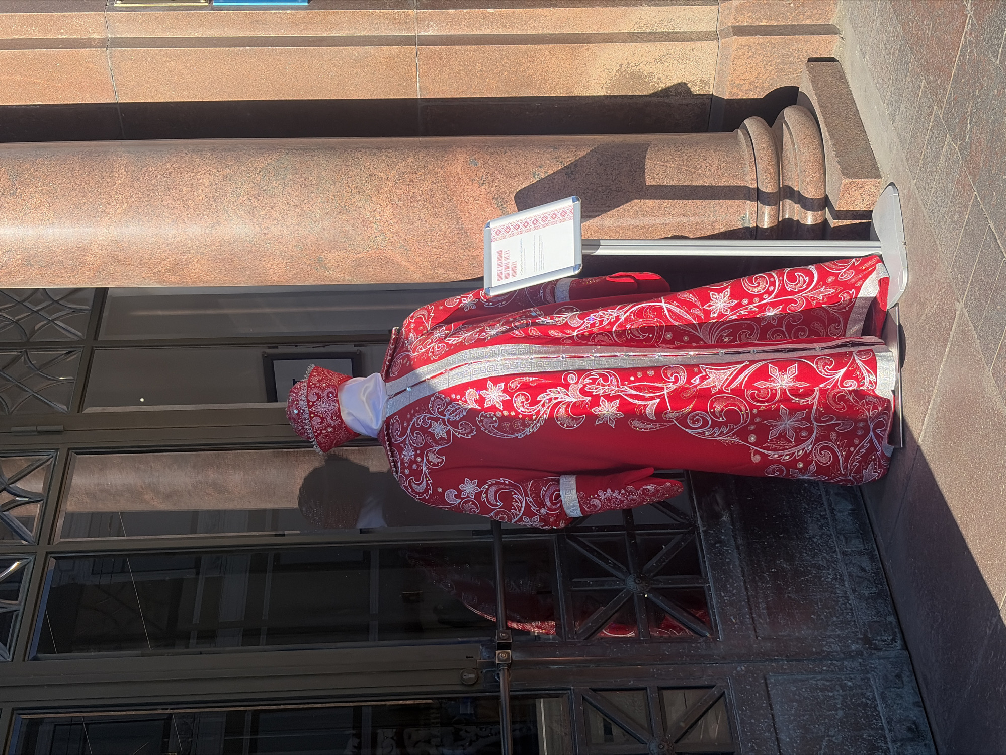 The image shows a mannequin dressed in an ornate red and white costume, standing in front of a building with large glass doors and a prominent reddish-brown granite column. The costume includes a long robe, a hat, and gloves, all decorated with intricate white embroidery and silver accents. A sign on a stand to the right of the mannequin displays text in Cyrillic script, suggesting the location is in Saint Petersburg, Russia, and likely related to an exhibition or cultural event. The sunlight casts strong shadows, indicating it is daytime. The ground is paved with square tiles.