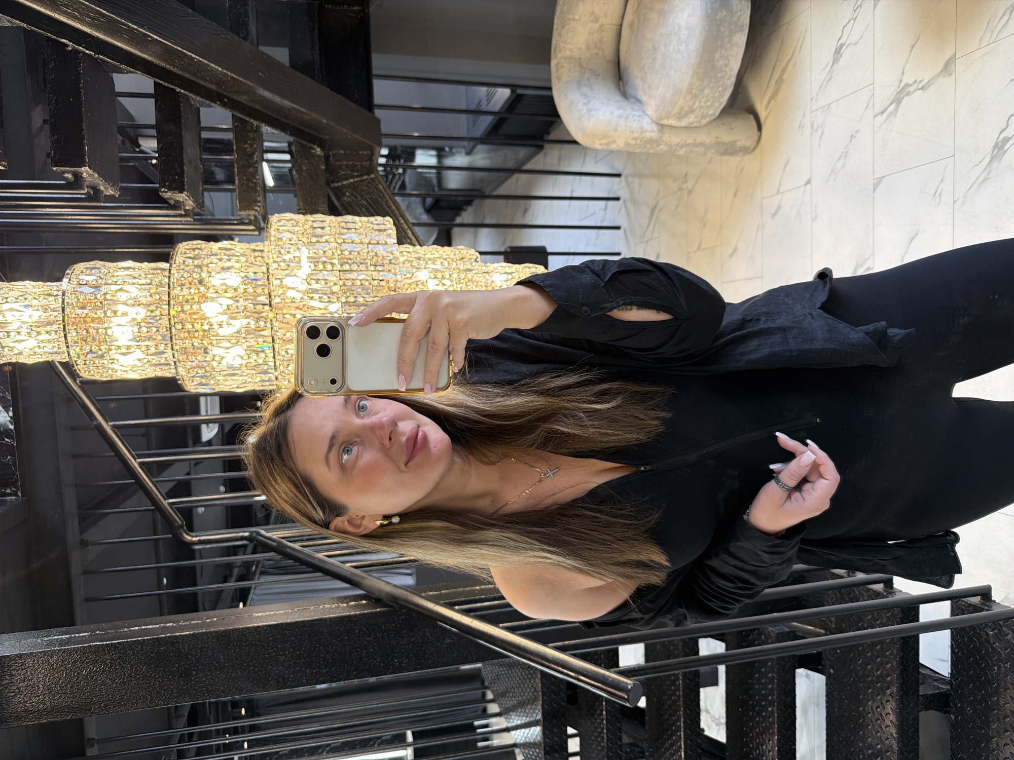 A woman is taking a mirror selfie in what appears to be a modern staircase and living area. She is standing near a black metal staircase with open risers, and a large, multi-tiered crystal chandelier hangs above and to her right. The floor is covered in large, marble-patterned tiles. To her right, a light gray, textured armchair is visible. The woman is wearing a black, off-the-shoulder top with long sleeves and a silver necklace with a cross pendant. She is also wearing a green ring on her right hand. Her hair is long and brown, and she is holding a smartphone in her right hand, angled to capture the reflection. The location is in the city of Укромновское сельское поселение, Ukraine.