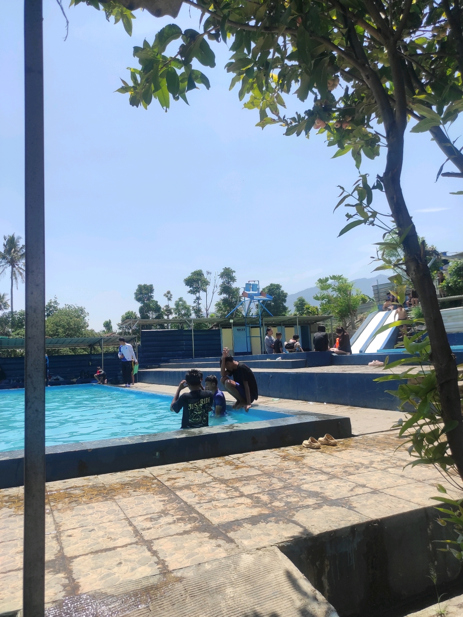 A sunny day at a water park in Ngamprah, Indonesia. A bright blue swimming pool occupies the majority of the image, with several people visible both in and around the water. In the foreground, two young boys are standing in the shallow end of the pool, one partially submerged. To their left, a young man in a black t-shirt with "JUST RIDE" printed on it is facing away from the camera, also in the pool. Further back in the pool, a group of people are gathered. On the right side of the image, a white and blue water slide is visible, with one person at the top and another at the bottom. People are seated on the edge of the pool and on benches in the background, some shaded by awnings. The environment is lush and green, with trees and mountains visible in the distance. The sky is clear and blue.
