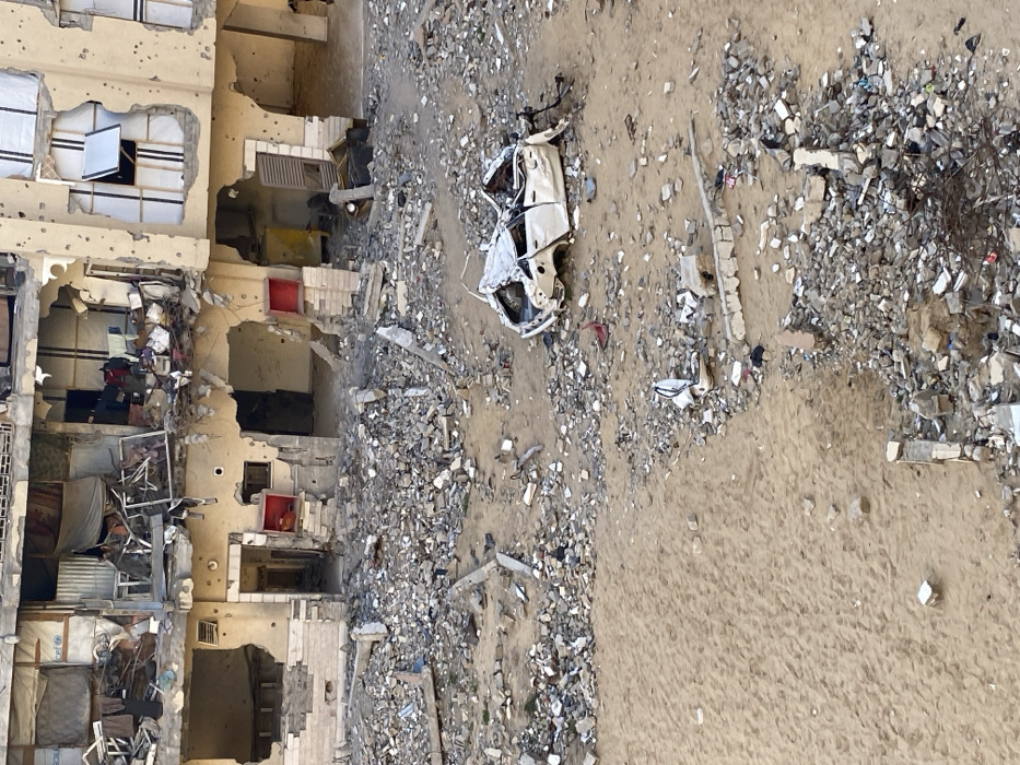 The image depicts a war-torn scene. A building, severely damaged with multiple floors exposed, stands adjacent to a sandy terrain littered with debris. A destroyed white car rests amidst the rubble, adding to the devastation. The building's facade is scarred with holes, revealing remnants of domestic life within. The overall impression is one of destruction and desolation.