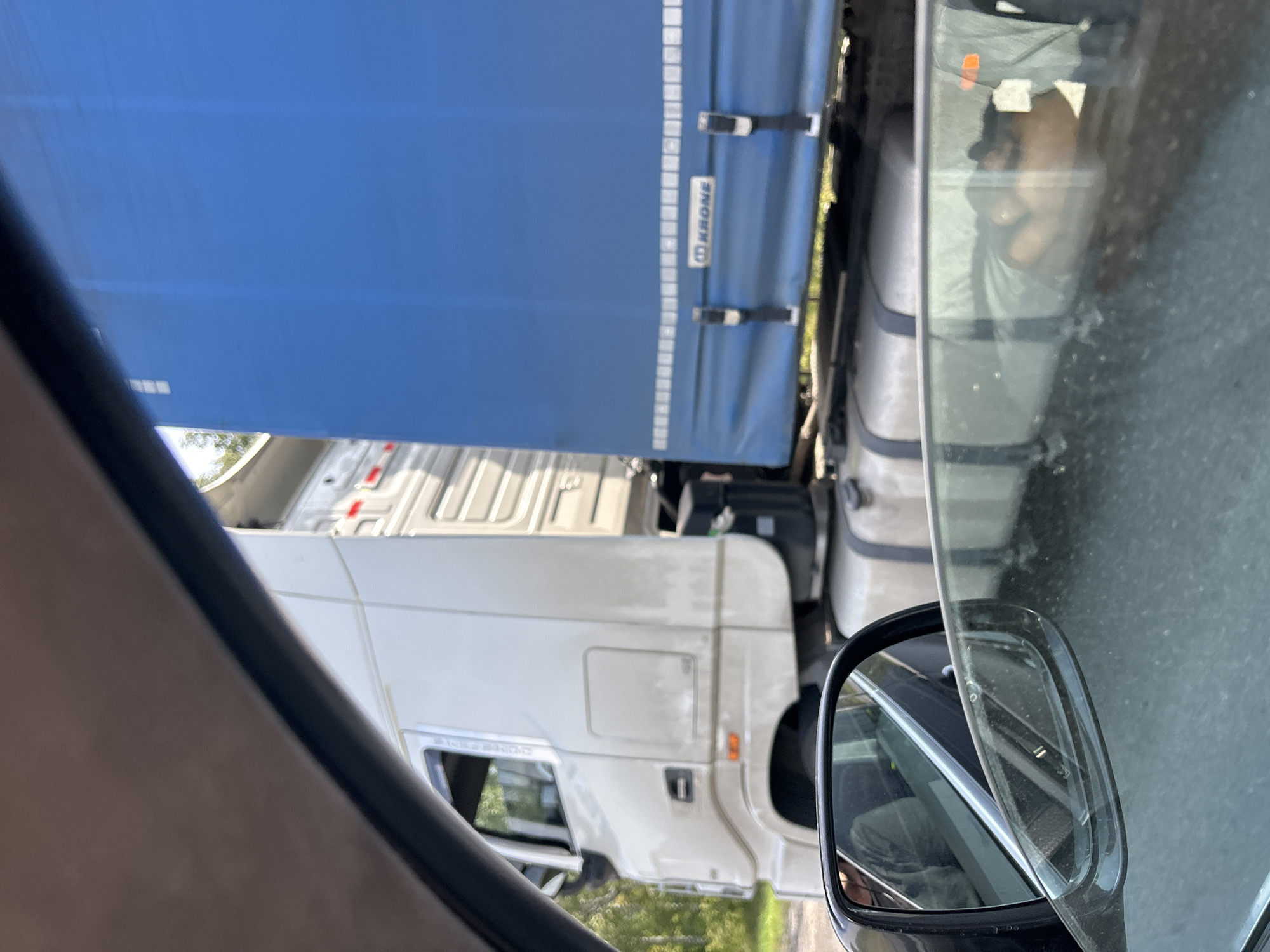 A white semi-truck with a blue tarp-covered trailer is parked on an unpaved road. The truck's cab is partially visible, and the trailer has the "KRONE" logo printed on it. Through the passenger side window of a car in the foreground, a person's arm and shoulder are visible, suggesting they are lying down in the vehicle. The background consists of trees and greenery, indicating a rural or roadside setting. The image appears to have been taken from inside a vehicle, looking out. The general location is identified as Spassk, Russia.