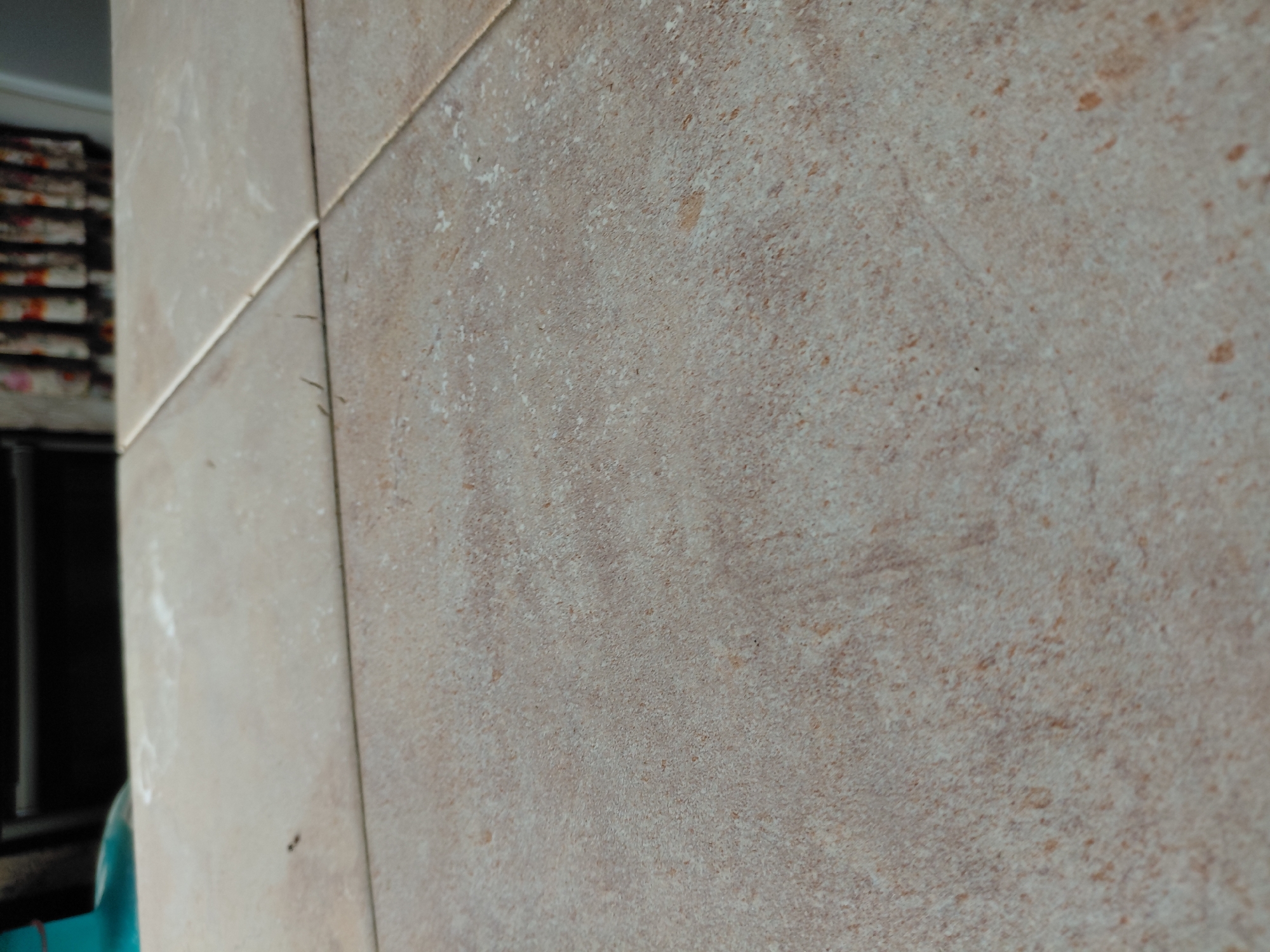 The image presents a close-up, angled view of a tiled surface, likely a wall or floor indoors. The tiles are square or rectangular, and their light beige to light brown coloration is accented by a speckled, textured pattern, suggesting a ceramic or porcelain material designed to mimic natural stone or concrete. Variations in color include lighter, grayish-beige areas and scattered darker, reddish-brown specks. Visible grout lines delineate the individual tiles, with one prominent horizontal and one prominent vertical line meeting near the center of the frame. The surface of the tiles appears to have a matte finish. Several small, white, irregular marks are present across the tiles, possibly dust, residue, or part of the inherent texture. In the blurry background on the left, stacks of rectangular objects, which could be books, papers, or patterned fabric, are visible. Below these, a dark, indistinct shape likely represents furniture or another structural element, and a portion of a bright turquoise object is faintly visible at the bottom left edge. There is no discernible activity taking place, and no people are visible. No text is present in the image. The time of day and weather cannot be determined from the visual information.
