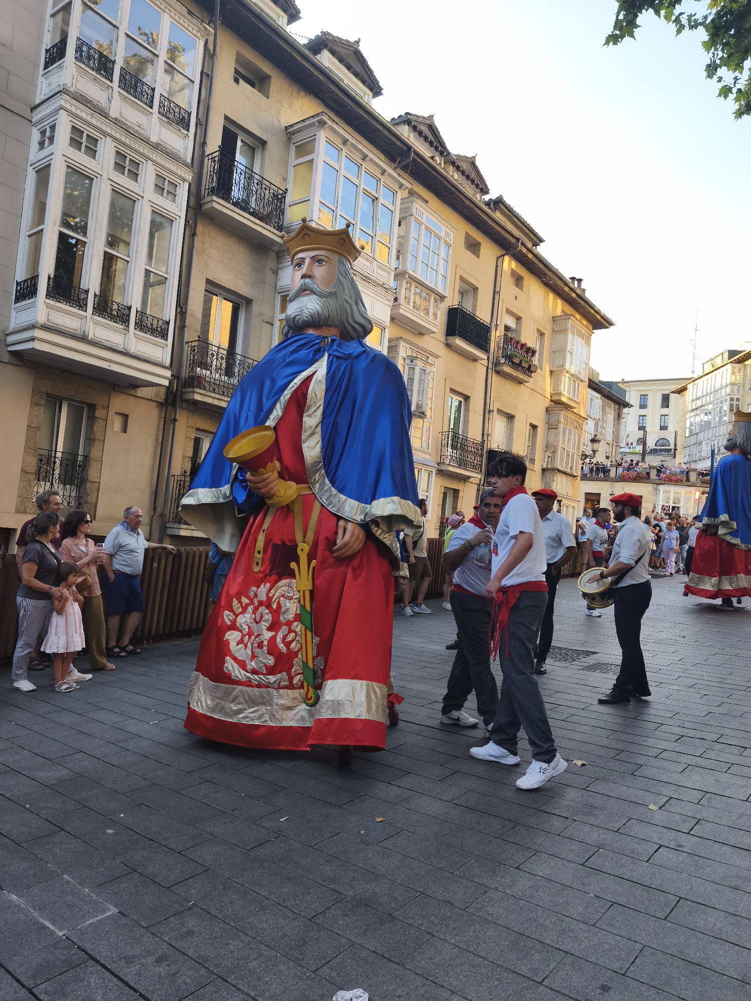 The image depicts a street scene in Vitoria-Gasteiz, Spain, likely during a festive event. In the foreground, a very large effigy of a crowned king, dressed in a blue cape and a red tunic adorned with intricate gold embroidery, stands prominently. The effigy holds a golden goblet in its left hand. To the left of the effigy, a group of onlookers, including adults and a young girl, are gathered, some appearing to interact with each other. To the right of the effigy, several individuals are engaged in activities, with one person playing a drum. Other large effigies are visible further down the street, suggesting a procession or parade. The background features traditional European architecture, characterized by buildings with multiple windows and balconies, illuminated by sunlight. The ground is paved with dark grey rectangular tiles. The sky above is clear and bright.