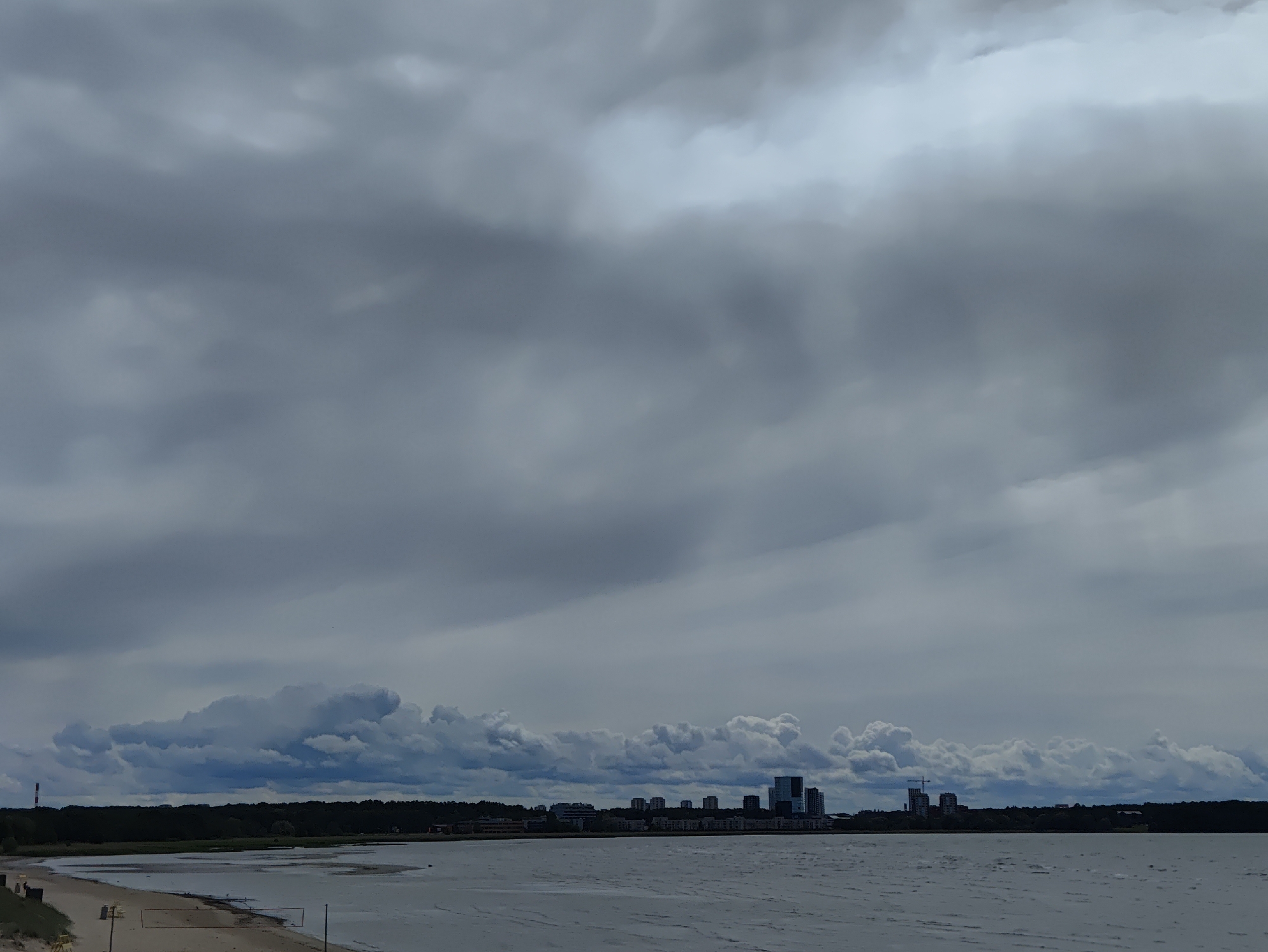 The image depicts a wide, overcast view of the Baltic Sea coastline near Tallinn, Estonia, with a city skyline in the distance. The sky is filled with heavy, grey clouds, suggesting an impending storm or a generally gloomy day. In the foreground, a sandy beach with volleyball nets is visible, indicating a recreational area. The water is choppy, reflecting the overcast sky. The middle ground features a line of dark green trees forming a dense forest along the coastline. Beyond the trees, the silhouette of Tallinn's modern skyline emerges, characterized by several high-rise buildings, including apartments and office blocks, and a construction crane. A tall, red and white industrial chimney stands out on the left side of the horizon. The scene conveys a sense of vastness and a somewhat somber, yet potentially active, urban and natural environment.