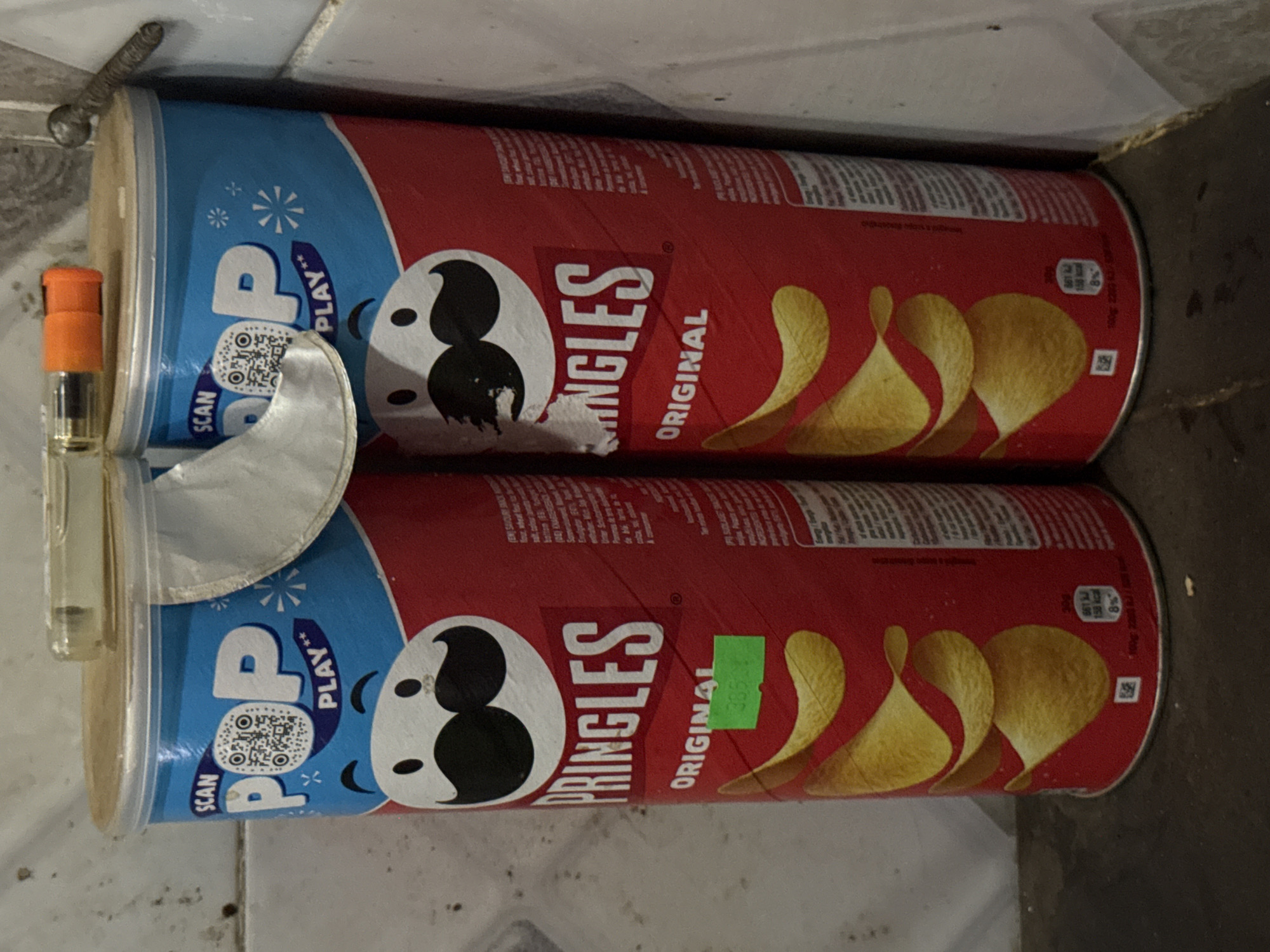 The image displays two Pringles "Original" flavor potato chip cans, horizontally stacked and resting on a dirty, light-colored tiled or concrete surface, leaning against a wall or corner.
The subjects include the two red and blue Pringles cans, both featuring the iconic Mr. Pringles character. A small, clear glass bottle with an orange cap, possibly a miniature perfume or spray bottle, is positioned vertically next to the top can. A piece of crumpled silvery foil, likely part of a Pringles seal, is visible near the bottle. A metal object, resembling a screw or bolt, is visible on the surface above the top can.
No activity or event is explicitly taking place; the items are stationary. The setting appears to be an indoor utilitarian space, possibly a floor or storage area, given the dirty surface.
Notable details include the packaging design of the Pringles cans, which feature "POP PLAY**" with a "SCAN" prompt and a QR code on the blue sections. A green price or identification sticker with the number "3850" is affixed to the lower can. The ambient lighting suggests an indoor environment.
Visible text on the cans includes "PRINGLES ORIGINAL", "POP PLAY**", "SCAN", and nutritional information such as "661 kJ / 158 kcal / 8%". Smaller text in English and German is also visible, mentioning "(EN) SAVOURY SNACK" and "(DE) KARTOFFELCESCHEN" (likely Kartoffelchips). The location context is Kurunduma, Nigeria.