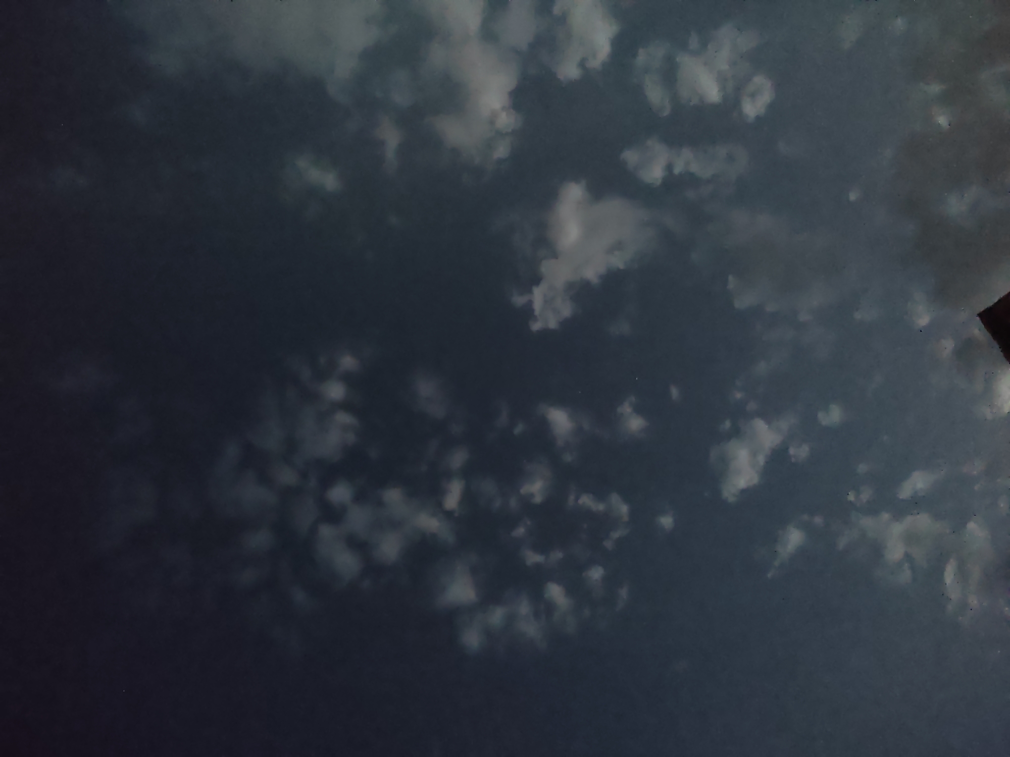 The image depicts a dark, cloudy sky, likely taken at dusk or night. There are scattered white clouds against a deep blue to near-black sky. A small, faint point of light, possibly a star or distant airplane, is visible in the upper left quadrant. The lower portion of the image shows the silhouette of a dark structure, possibly a rooftop or the top of a building, with a triangular peak. The scene is devoid of any visible people or specific landmarks that would pinpoint a precise location within Арчалы, Kyrgyzstan. The overall impression is one of a quiet, possibly overcast evening sky over a sparsely populated area.