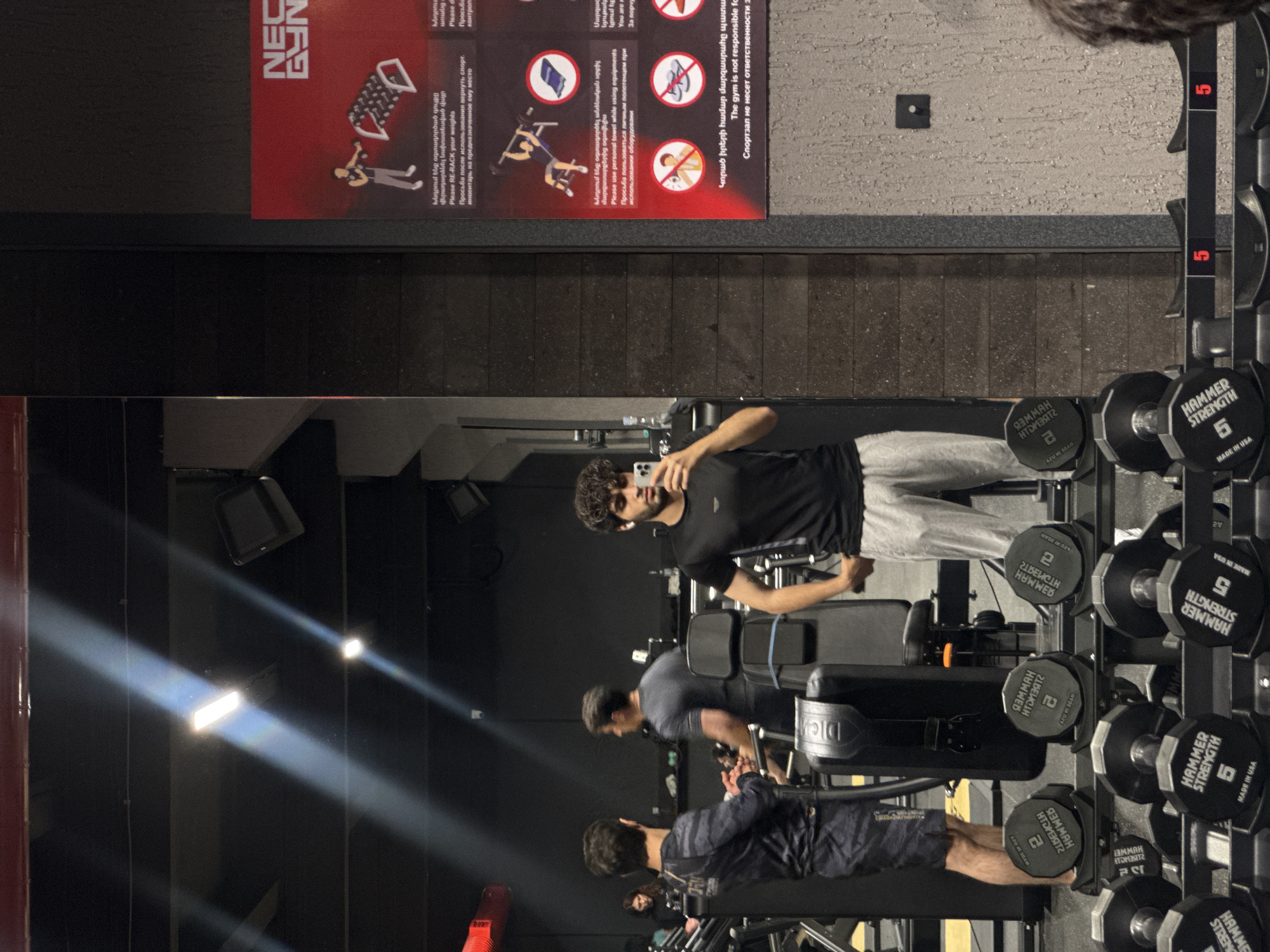 The image depicts the interior of a gym. In the foreground, a young man with curly hair is standing, holding a smartphone in his right hand and looking at it. He is wearing a black t-shirt and light gray sweatpants. Behind him, to the left, another man in a dark patterned long-sleeved shirt and dark shorts is facing away from the camera, talking to someone not fully visible. Further in the background, several gym machines and racks of dumbbells are present. On the right side of the image, a red poster with various instructional images and text in Armenian and English is attached to the wall. The poster contains rules and information about the gym, including warnings and equipment usage guidelines. The overall environment appears to be a modern gym with artificial lighting. The presence of the poster with Armenian text strongly suggests the location is in Yerevan, Armenia.