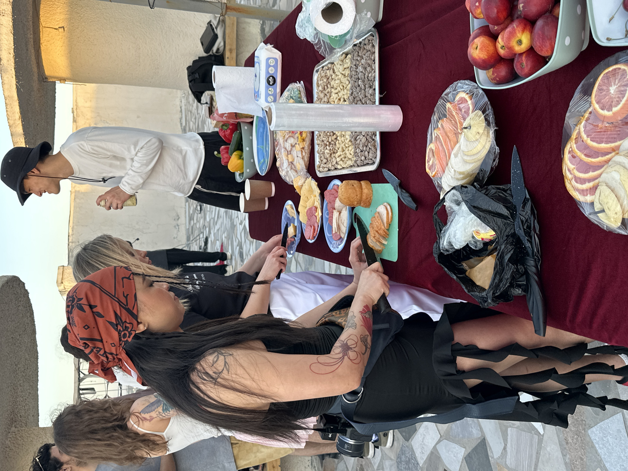 The image shows a rooftop gathering in Tashkent, Uzbekistan. A table covered with a maroon cloth is laden with food: peaches in a green container, sliced oranges, nuts in metal trays, sliced meats on plates, and bread. A man in a white shirt and black hat stands at the end of the table, holding a can. Several people are gathered around the table, some holding knives and preparing food. A woman with a patterned bandana is prominent in the foreground. A black trash bag sits on the table. The background shows a light-colored wall and a tiled surface.