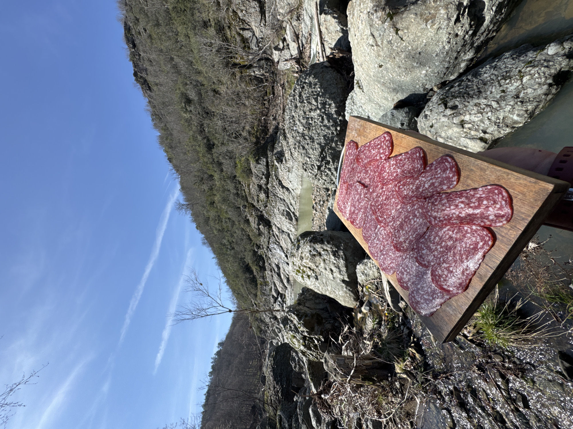 The image captures an outdoor scene within a rocky river valley in Fazanovo, Bulgaria, under a clear blue sky marked by faint contrails. In the lower right foreground, a partially visible human hand, wearing a dark wristband, holds a rectangular wooden cutting board. The board is covered with multiple overlapping, thinly sliced pieces of red salami, showing distinct white marbling. To the immediate left of the board, a narrow stream flows over dark, wet rocks and sparse green vegetation. The surrounding environment consists of a riverbed dominated by large, irregularly shaped grey boulders. Sections of still, light-green tinted water are visible among the rocks, indicating a shallow river or creek. Sparse, bare-branched trees and shrubs are scattered throughout the riverbed and along its banks. A solitary, slender, leafless tree stands prominently in the midground. Steep hillsides, densely covered with mostly bare deciduous trees and some evergreen shrubs, frame the valley in the background under bright, clear sunlight.