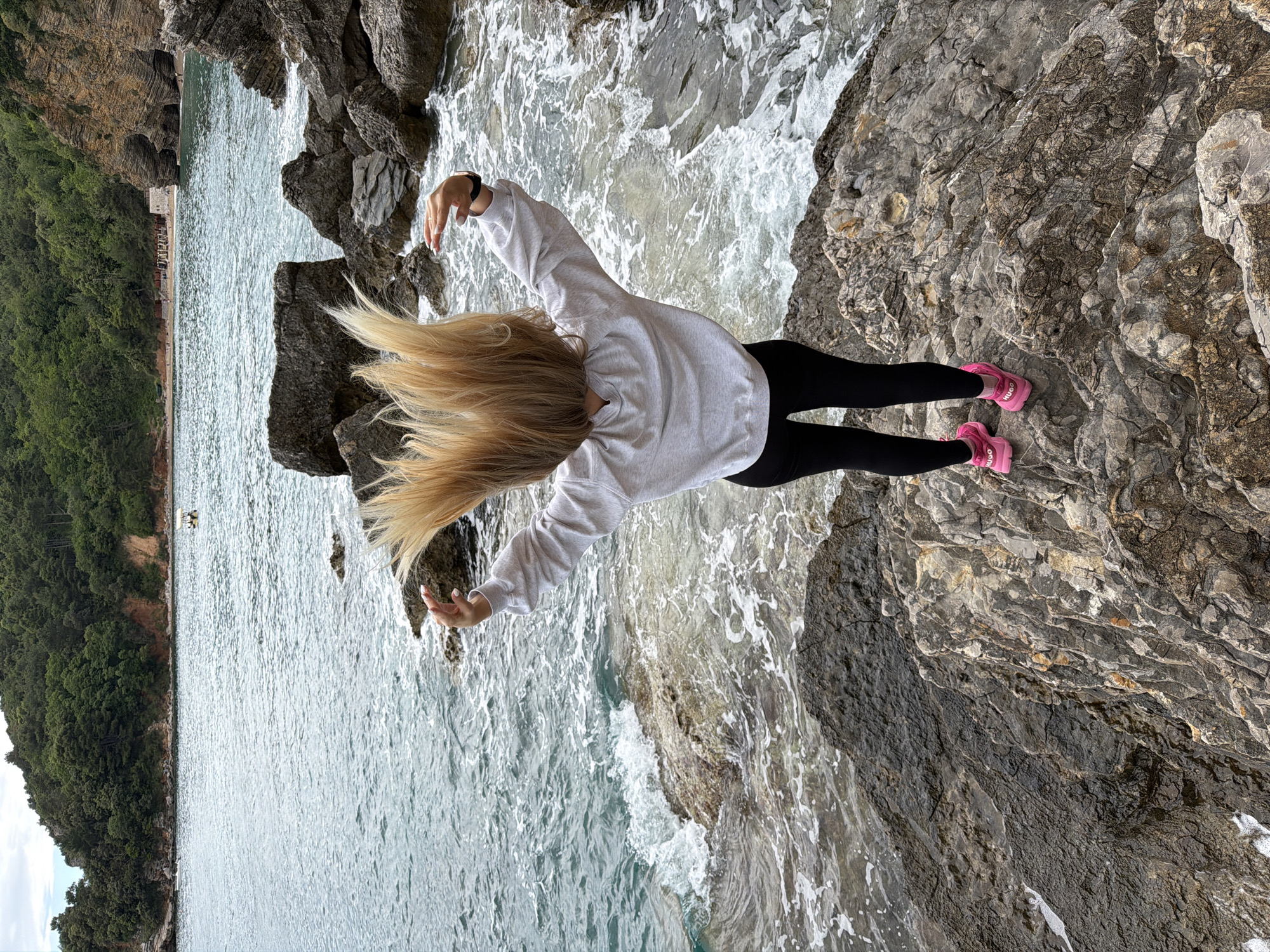 A person with long blonde hair, wearing a gray sweatshirt, black leggings, and pink sneakers, stands on a rocky cliff overlooking the Adriatic Sea in Budva, Montenegro. The person's arms are outstretched, and their hair is flowing forward. The sea is visible on both sides of the rock formation. Vegetation covers the cliffside.