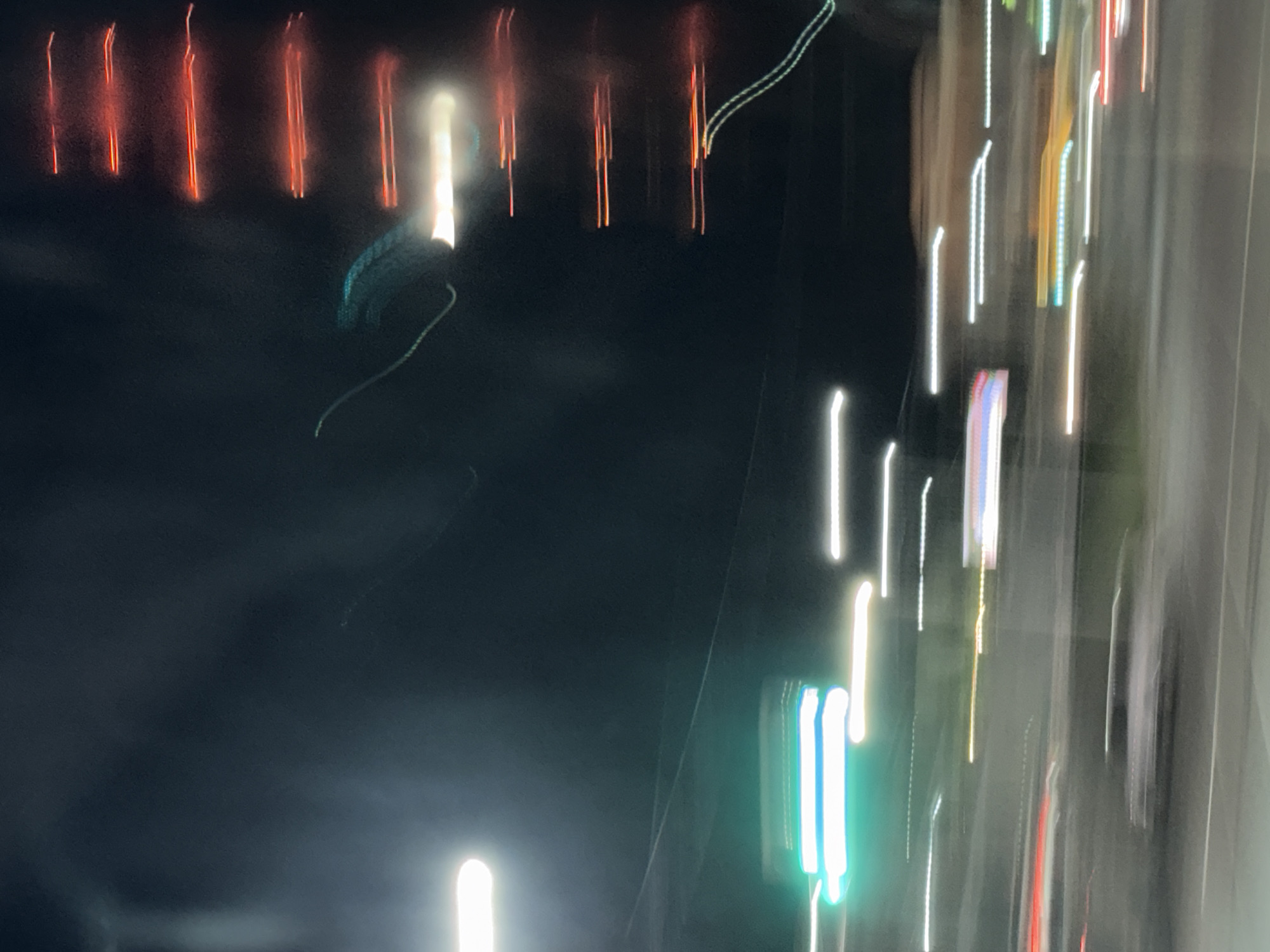 The image is a blurry long exposure shot taken at night in an urban area of Ufa, Russia. Red and orange vertical light streaks are visible at the top left, likely from building lights or traffic signals. To the right and lower are bright white, green, and blue vertical light streaks from unidentified light sources, possibly headlights or streetlights. A faint snaking light trail is also present. The overall scene conveys a sense of motion and urban activity.