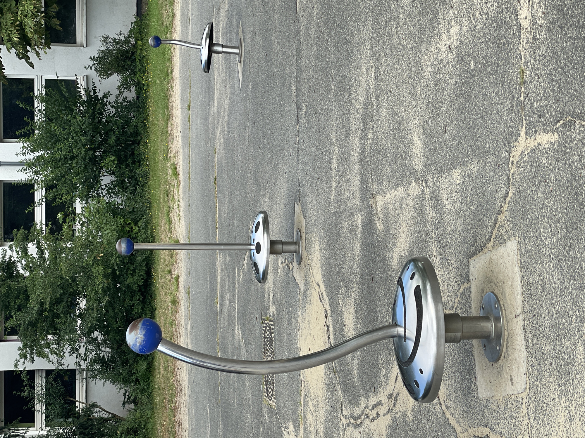 The image depicts a park-like area with a building in the background and three outdoor exercise or play structures in the foreground. The structures are made of polished metal and consist of a circular base with holes, a vertical pole, and a curved arm topped with a blue sphere. The ground is paved with asphalt and has some sandy patches and cracks. The building in the background appears to be an institutional building with multiple windows. The overall setting suggests an outdoor public space in Bremerhaven, Germany.