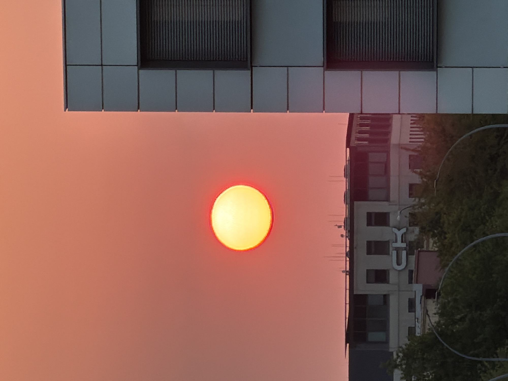 The image captures a sunset in Leudelange, Luxembourg. A bright, yellow sun with a red halo is prominently positioned in the center-left of the frame, set against a soft, orange sky. To the right, a modern, grey-tiled building with horizontal blinds covering its windows occupies the right side of the image. In the background, a lower, lighter-colored building with visible windows and the letters "CK" in large white signage is partially obscured by dark green trees. Streetlights and lampposts are also visible in the foreground and mid-ground. The overall scene depicts an urban landscape at dusk.