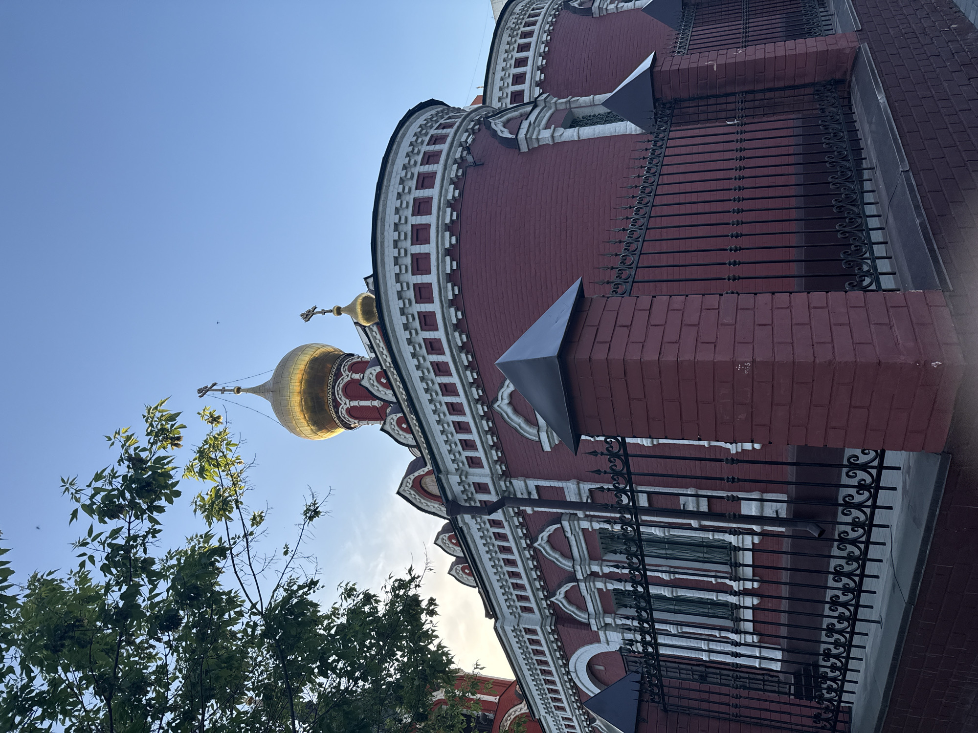 The image shows an upward angle view of a red brick building with a golden dome and cross, likely an Orthodox church. The architecture features white decorative elements and a dark metal fence in the foreground. Green trees with leaves partially obscure the left side of the building and dome. The sky is a clear, light blue. The location is likely in Moscow, Russia, given the architectural style of the church.