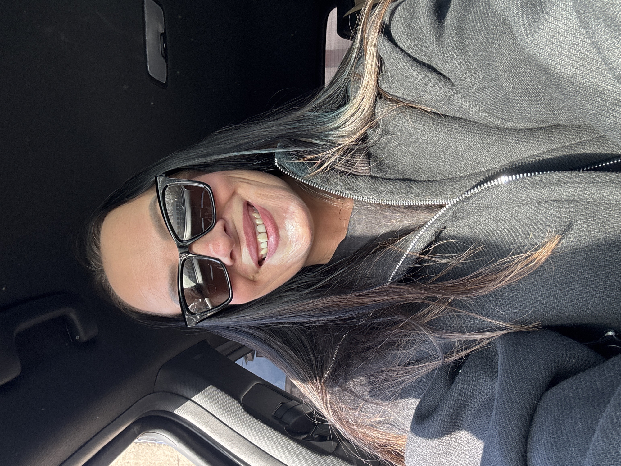 An adult female is depicted taking a selfie from the driver's seat of a vehicle in Chaikovsky, Russia. She is smiling broadly, exposing her teeth, and wears dark-framed sunglasses. Her long, dark hair features lighter brown and subtle blue-green highlights. She is dressed in a dark gray zippered jacket over a dark t-shirt. The vehicle's interior includes a black headliner, a visible grab handle, and a dark A-pillar on the left. Through the driver's side window, a blurred outdoor environment is partially visible, showing a light-colored building structure and bare tree branches against a bright sky, indicating daylight conditions. Reflections of the exterior are present on the sunglasses. No other individuals or significant actions are observed.