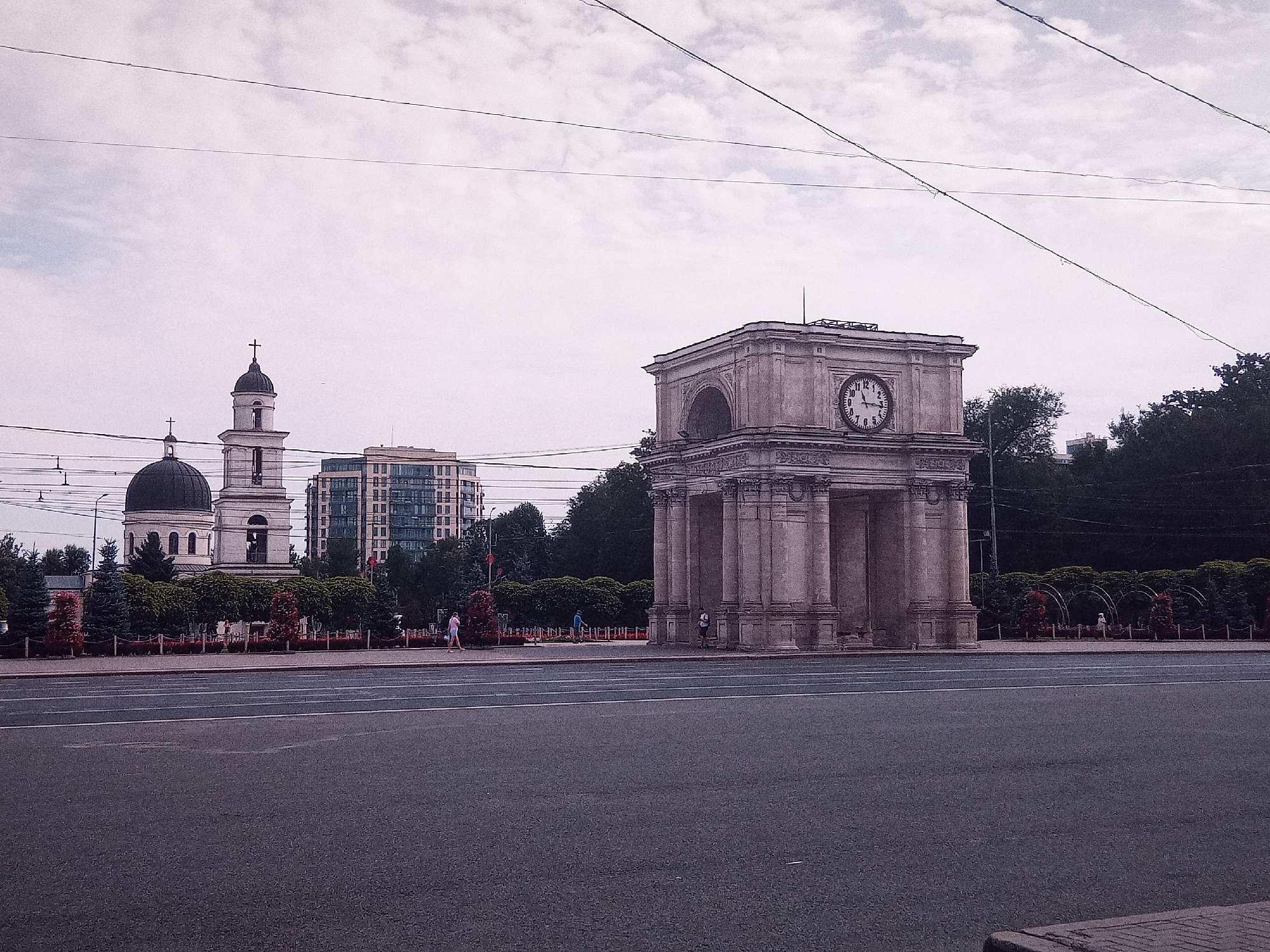 In Chișinău, Moldova, the view captures a street scene with the Triumphal Arch on the right and the Nativity Cathedral in the background on the left. The Triumphal Arch, a neoclassical monument, features a prominent clock face and large stone columns. The Nativity Cathedral, an Orthodox church, displays a distinctive dark dome and a tall bell tower topped with a cross. Adjacent to the cathedral, a modern glass-fronted building rises. The foreground consists of a wide asphalt road with white lane markings. A paved walkway with a low hedge and decorative trees separates the road from the monuments. Several individuals are visible on the walkway: a person in a pink top and shorts walks to the right, another person in a blue shirt walks left, and a person in white clothing stands near the arch. Power lines crisscross the overcast sky. The overall atmosphere is calm and urban.