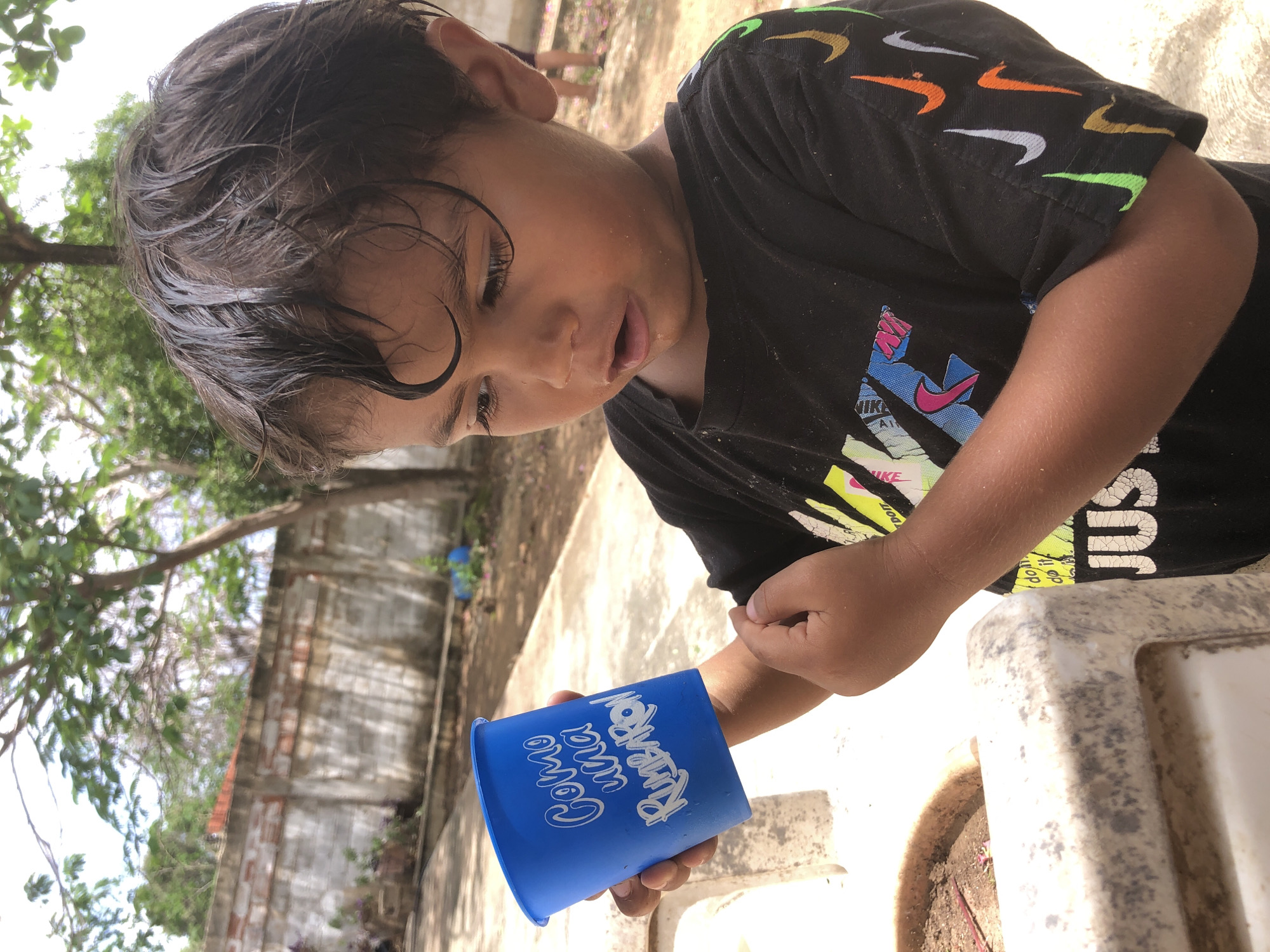 A young boy with wet, dark hair is looking down at a blue plastic cup in his hand. He is wearing a black t-shirt with a pattern of colorful Nike swooshes on the sleeves and a graphic on the front. The boy's face is partially obscured by his hair and he appears to be concentrating on something in the cup. The background shows a rough, outdoor setting with a concrete floor, a brick wall, and trees with green foliage. The scene is outdoors, likely during the day given the bright lighting. The blue cup has white text on it, which is partially visible and appears to say "Como un" followed by more text that is difficult to read. The overall impression is of a child engaged in play or exploration in a casual outdoor environment.