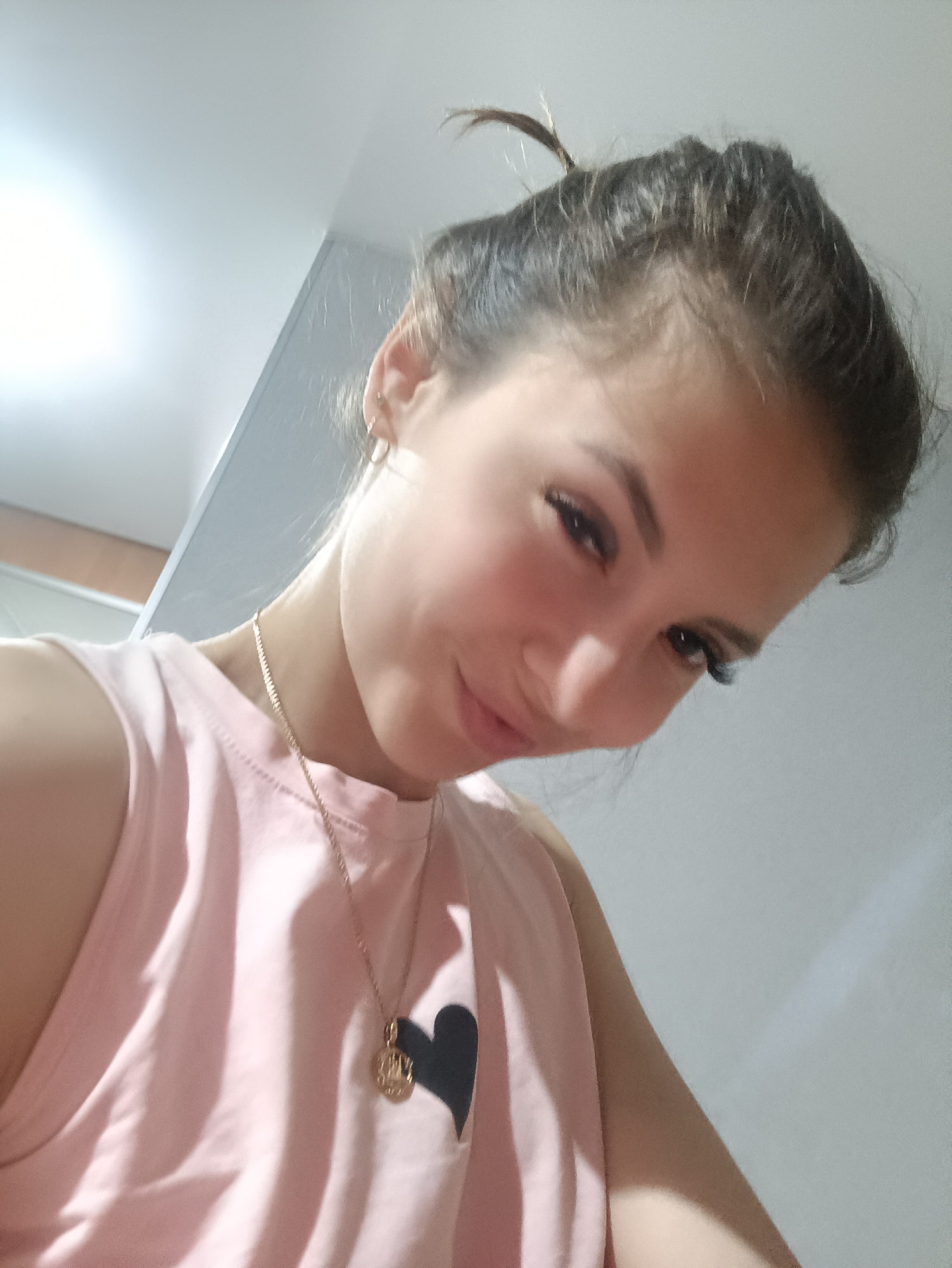 A young woman is visible in a close-up photograph. She has her brown hair pulled back in a bun, with a few strands loose on top. She wears a light pink sleeveless shirt with a black heart graphic, and a gold necklace with a round pendant. Her expression includes a slight smile while she looks downward. She has fair skin. The background appears to be an indoor space with plain, light-colored walls and a ceiling. The location details are unknown.