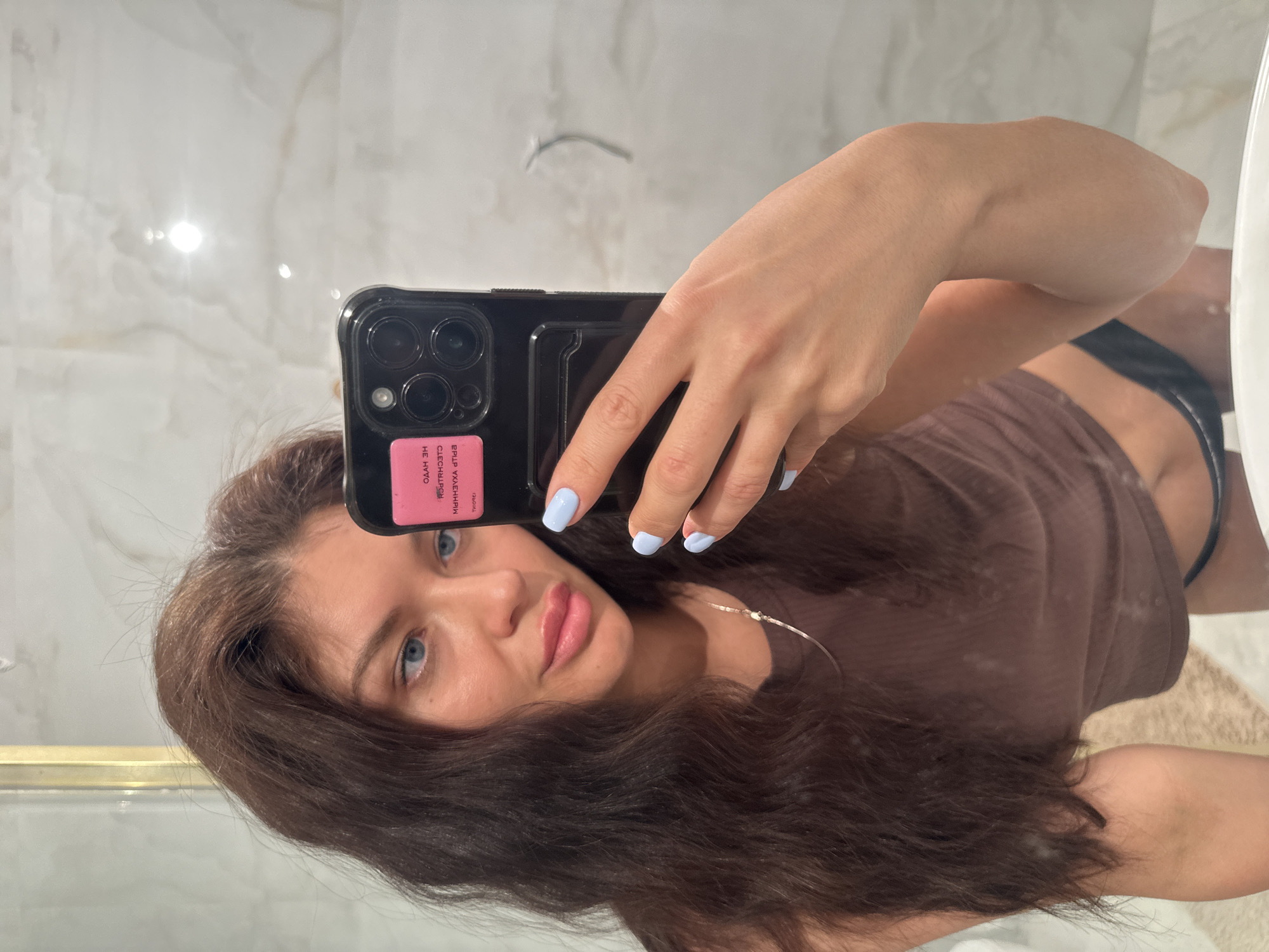 A woman takes a selfie in a bathroom. The location is most likely not in Khimki, Russia as there is nothing in the image that suggests this location. The woman is holding a black smartphone with a pink sticker on the back in her right hand. She is wearing a brown top and her hair is dark brown. Her nails are painted light blue. The bathroom features a white marble wall and a sink.