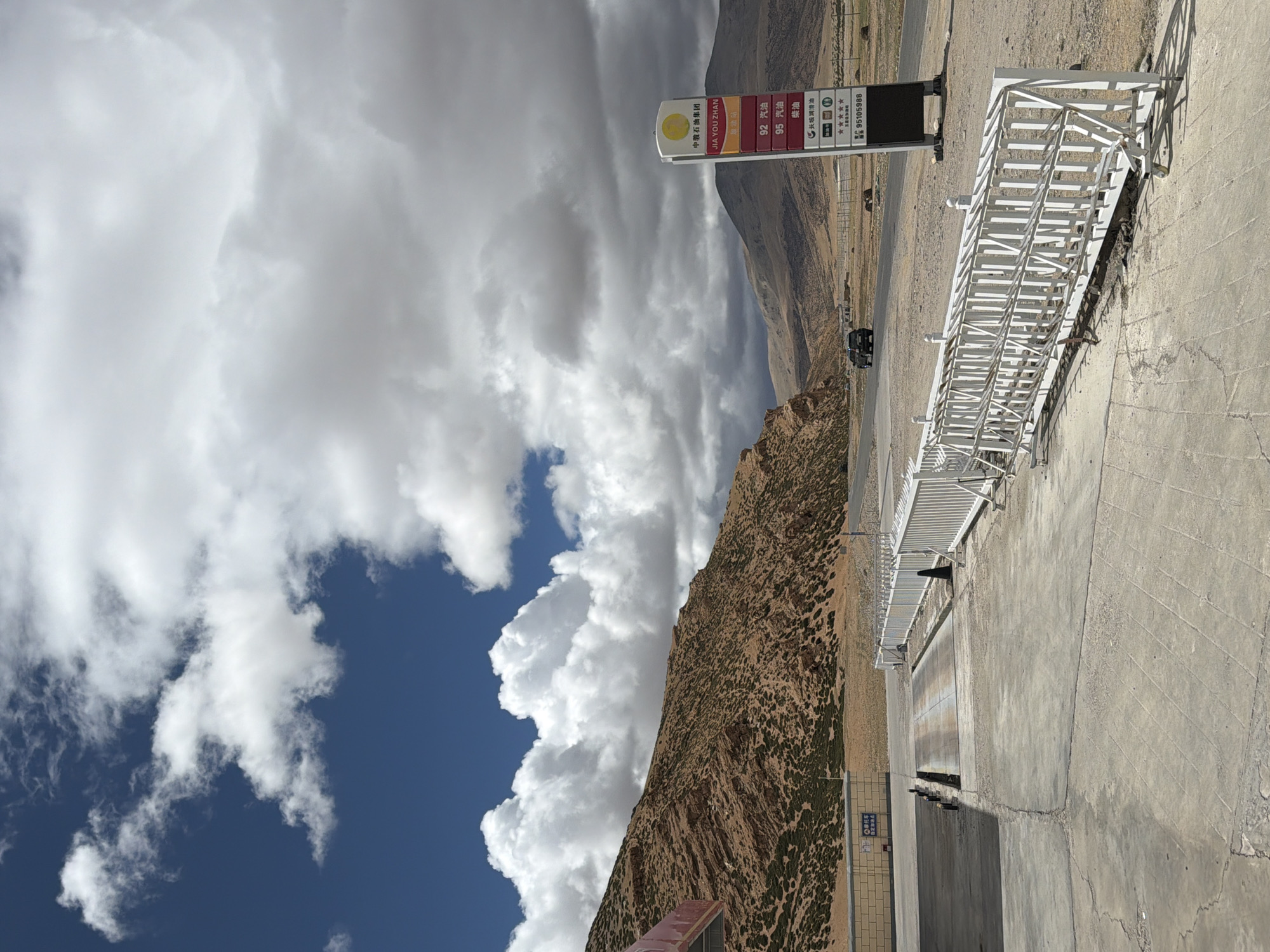 The image captures a scene in Kela Township, China, featuring a petrol station sign displaying fuel prices, the name "JIA YOU ZHAN" and "中骏石油集团," a mountain backdrop, a white metal structure that could be part of a fence or barrier, a vehicle on the road, and a partly cloudy sky. The ground is concrete, suggesting an industrial or commercial area. The sign also lists a phone number and the brand of oil "长城润滑油".