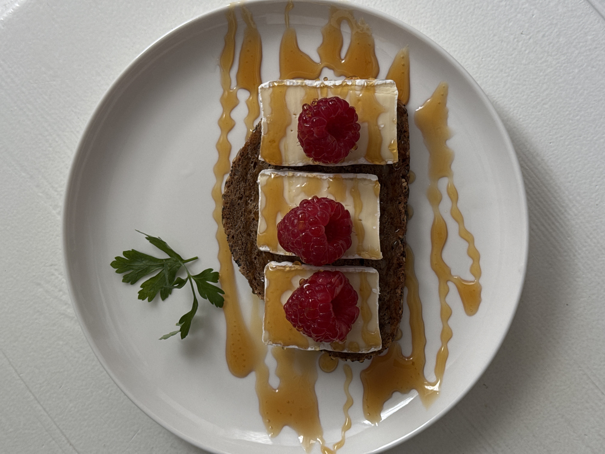 A close-up, top-down shot shows a piece of dark bread topped with three pieces of white cheese, each adorned with a single raspberry. A drizzle of honey is artfully spread across the cheese, bread, and the white plate. A sprig of parsley sits to the left of the food. The image appears to be taken indoors, possibly in a kitchen or dining area, on a white table. The food is the central focus, presented in a neat and appetizing manner. There are no people visible in the image. The setting does not provide any specific clues to its location within Siegburg, Germany.