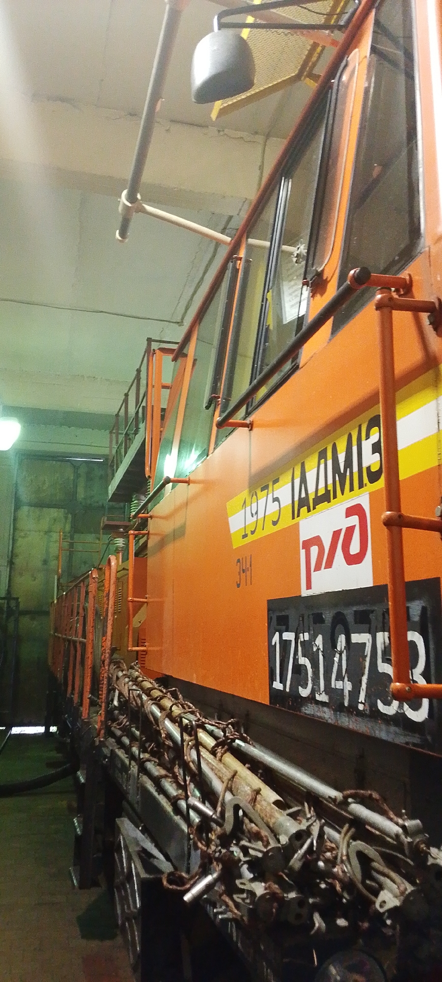 In Bira, Russia, inside what appears to be a train depot or maintenance facility, an orange train car is prominently featured. The car has visible markings, including the number "1975", the word "ЛАДМИЗ", and the logo of RZD (Russian Railways). Below these markings, the number "341" is present, and further down, a large serial number "17514753" is displayed in white on a black background. The train car is equipped with an orange railing system and a side ladder. The foreground is dominated by a collection of metal rods and pipes, bound together with rope, resting on a lower section of the train car. The environment is dimly lit, with overhead lighting visible, and the walls appear to be made of concrete or a similar industrial material. The overall impression is one of an industrial setting, likely a railway maintenance or storage area.