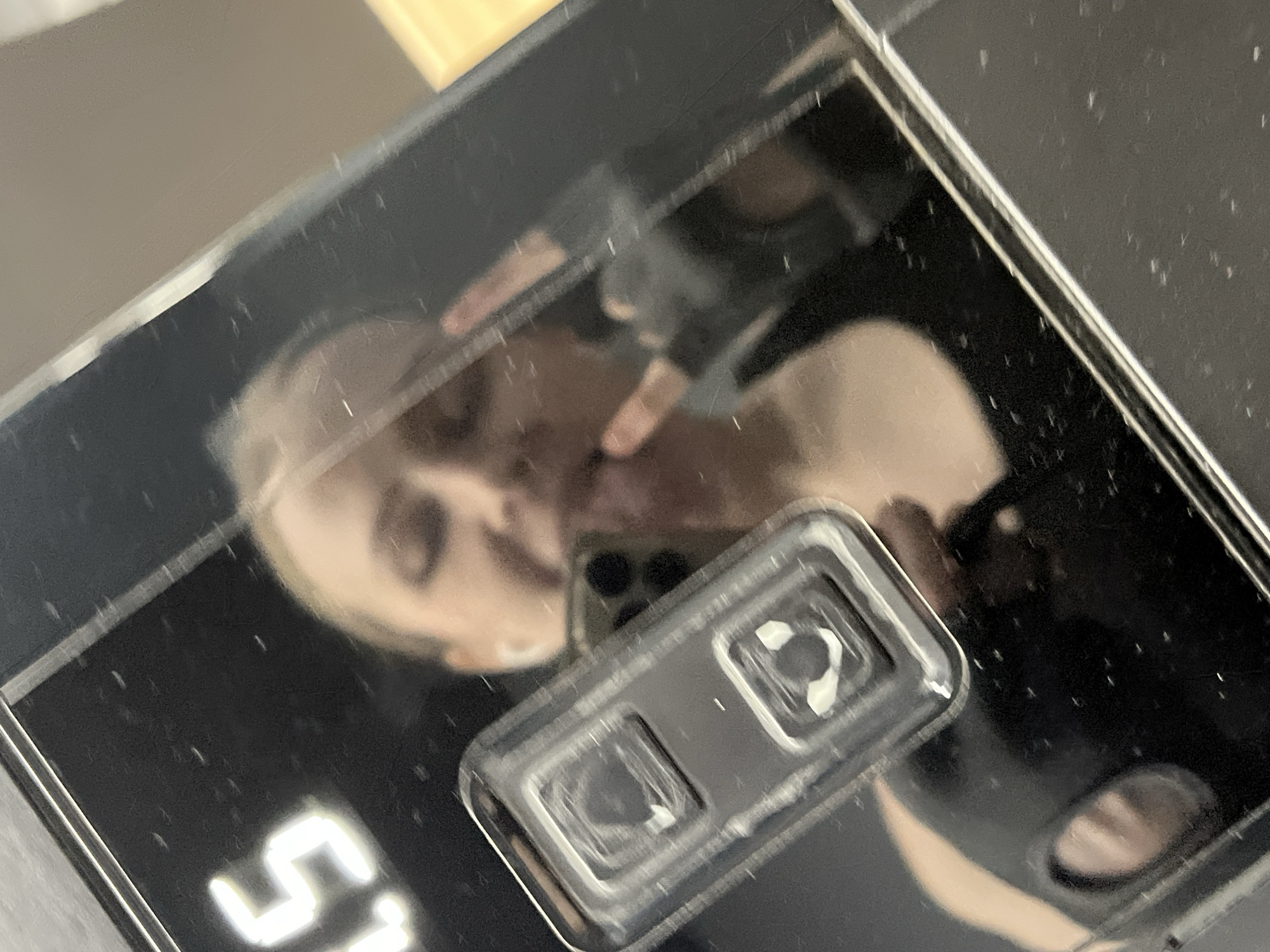 This close-up image displays a blurred reflection on a dark, glossy surface, likely glass or highly polished material, which is marked with numerous small scratches or dust particles. A person with light-colored hair is reflected, looking downwards towards the surface or the device taking the photo. Their face is partially visible and out of focus, showing glimpses of an eye and eyebrow.
Prominently reflected near the center-right of the image is a dual camera module, suggesting the presence of a smartphone or similar device. Towards the bottom-left of the reflective surface, a bright, glowing white number "5" is visible, accompanied by what appears to be part of another digit or character. The reflected background is indistinct but hints at an indoor setting, with some lighter areas possibly indicating a wall or wooden surface. The scene captures a moment of self-reflection or interaction with a device, possibly examining the glossy surface. The location context for this image is Khimki, Russia.