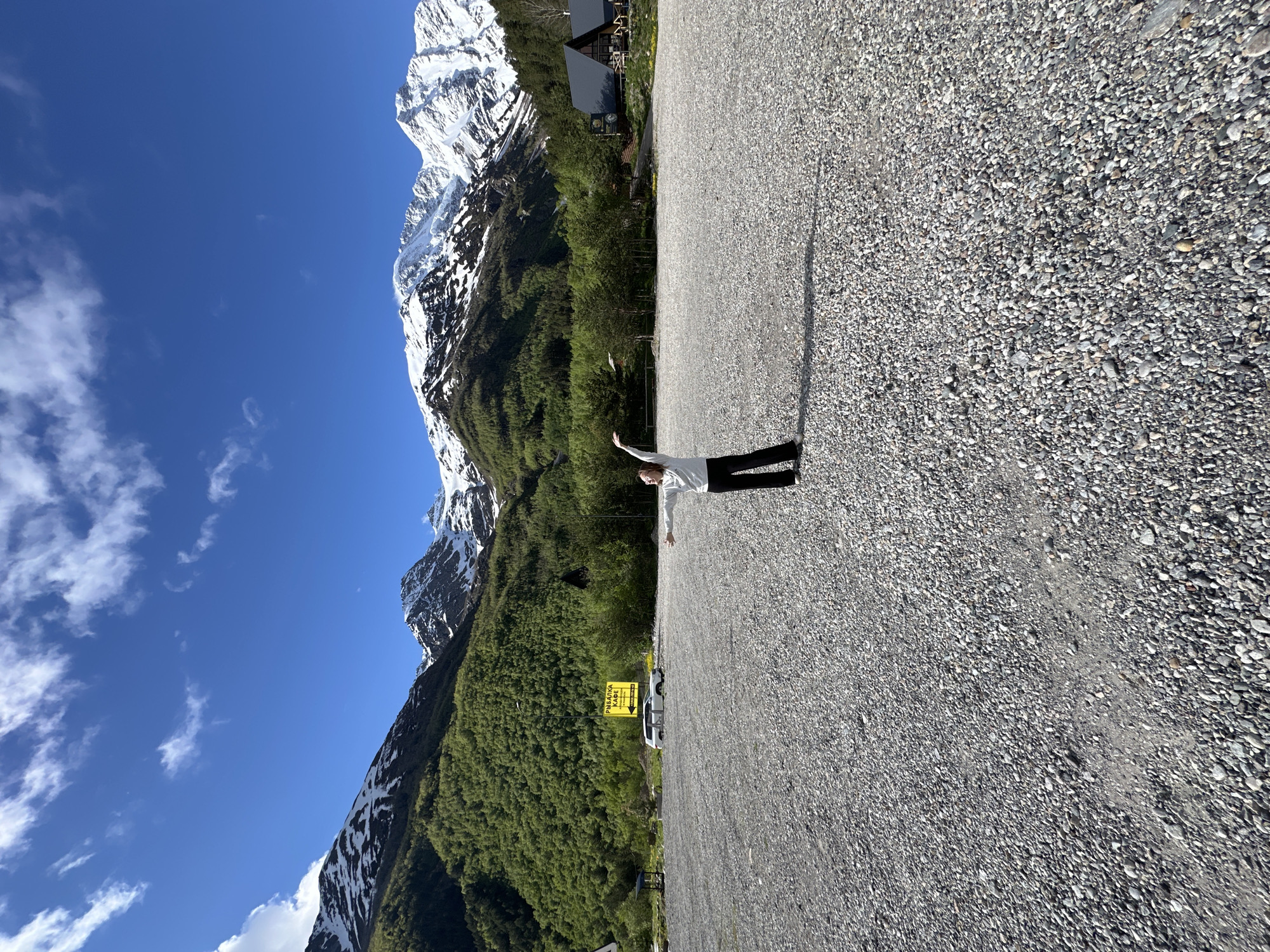 In Terskol, Russia, a young person with blonde hair, wearing a light-colored sweatshirt and dark pants, stands in a vast, open area covered with grey pebbles. They are positioned in the center of the frame, facing the viewer with their arms outstretched to the sides, as if in a welcoming gesture or mid-dance. A long shadow is cast behind them on the pebbled ground, indicating a sunny day.
The background is dominated by the impressive natural landscape of the Caucasus Mountains. Lush green trees cover the lower slopes of the mountains, transitioning to snow-capped peaks under a vibrant blue sky dotted with white, wispy clouds.
To the left, a yellow sign with Cyrillic text, which appears to read "РЫБАЛКА КАФЕ" (Fishing Cafe), is visible, along with a white vehicle parked nearby. Further to the right, nestled amongst the trees at the base of the mountains, are several dark-colored, angular buildings that resemble cabins or small houses, some with visible windows and dark roofs. The overall scene depicts a serene, picturesque mountain setting with signs of human presence and activity.