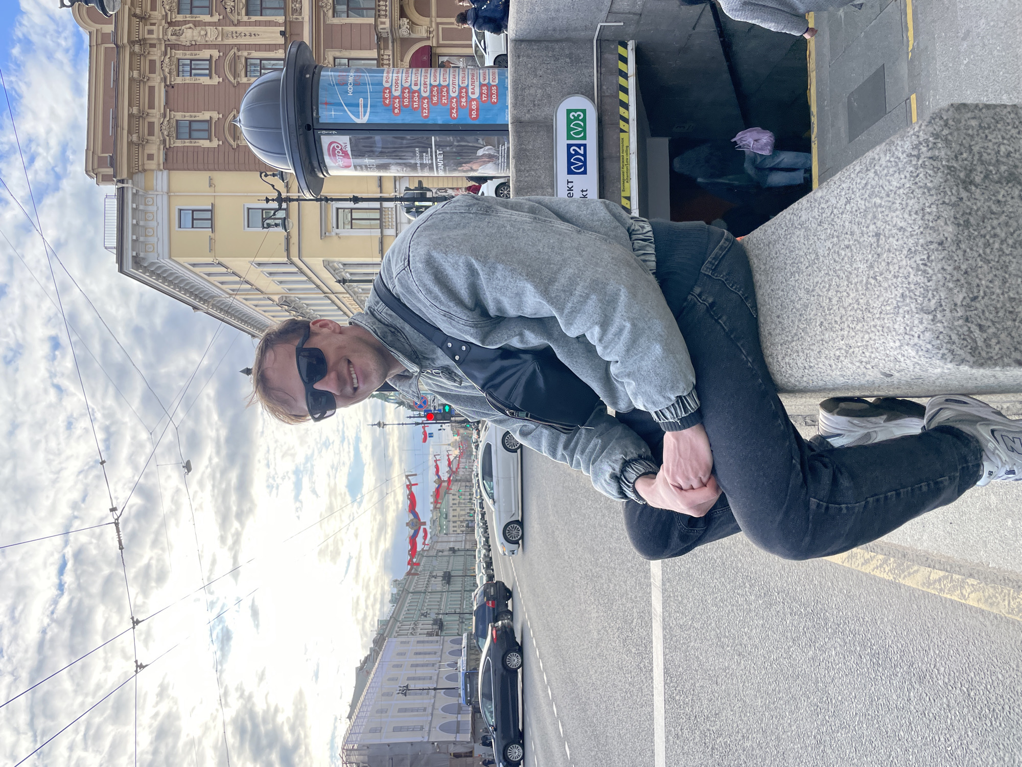 A young man is captured in a daylight photo, smiling directly at the camera while sitting on a grey, textured stone barrier. He wears a grey denim jacket, a black bag across his shoulder, black jeans, and sunglasses, with his hands clasped in front of him.
The scene is an urban street in Saint Petersburg, Russia. In the background to the left, ornate, multi-story buildings with classical architecture line the street under a bright, cloudy sky. Overhead wires, possibly for trams or trolleys, crisscross the upper portion of the frame.
To the right of the man, a metro station entrance is visible, marked by a sign with "V2" (blue circle) and "V3" (green circle) indicating metro lines, and Cyrillic text including "метро" (metro) and "ект" (likely part of "проспект"). An advertising column stands prominently, displaying various event listings and dates in Cyrillic, such as "КОСМОРО," "ОПИЯ ВЫСОЦКАЯ," "ГАМЛЕТ," and specific dates like "4.04" and "9.04." An informational sign at the metro entrance warns "ВНИМАНИЕ Низкий проход" (Attention Low headroom) and "Attention Low ceiling."
A street with parked and moving cars is in the midground, with red and white decorative banners visible further down the street. The overall impression is a bustling city environment.