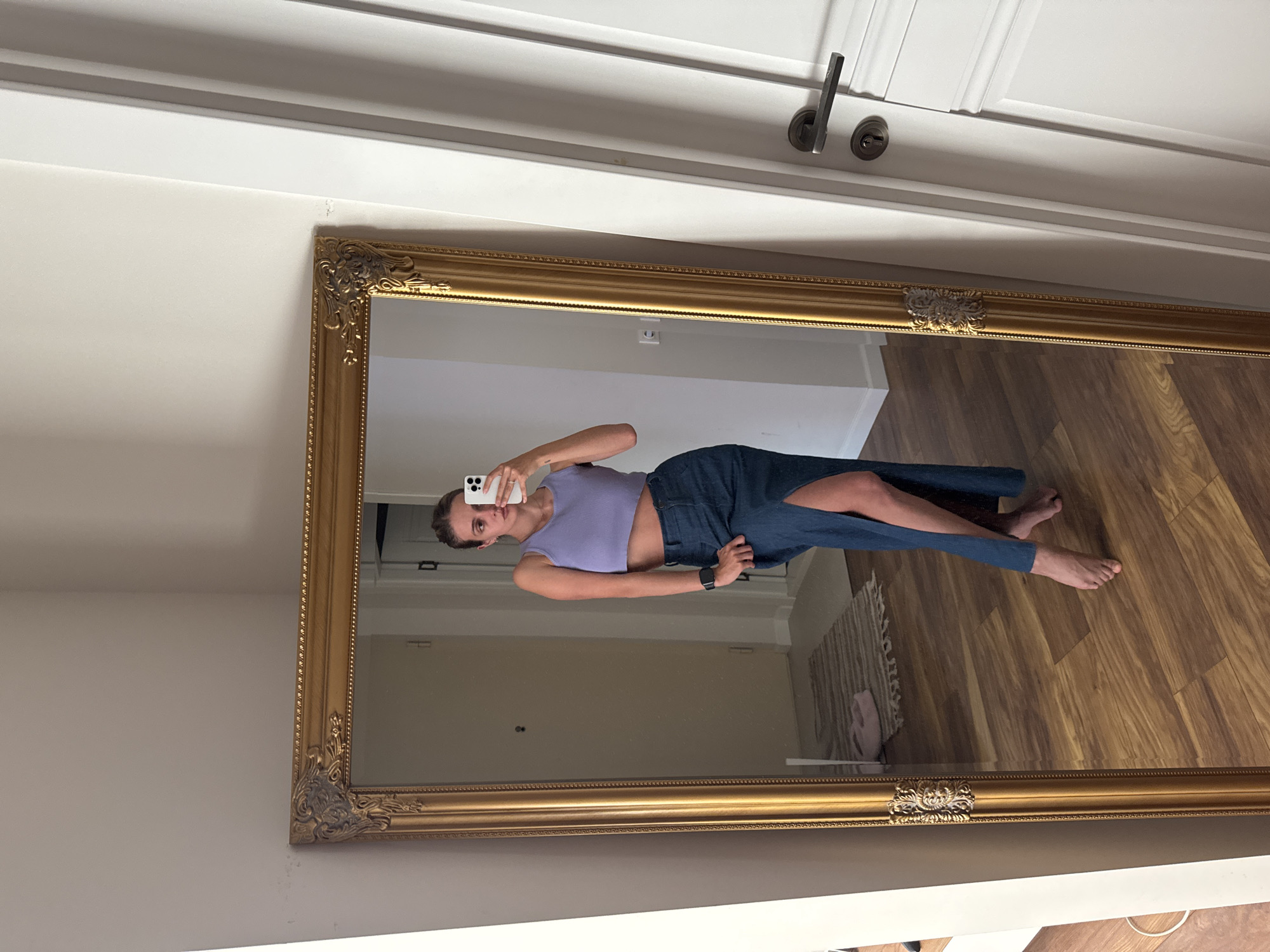 This media file captures a mirror selfie, rotated 90 degrees counter-clockwise from a standard upright orientation. The central subject is a person, appearing to be a young adult, reflected in a large, ornate gold-framed full-length mirror.
The person is wearing a light purple sleeveless crop top and a long, dark denim skirt with a high slit on the side, revealing one bare leg. They are holding a white smartphone, which covers part of their face, as they take the mirror selfie. Their other hand rests on their hip. Their bare feet are visible at the bottom of the reflection.
The reflection within the mirror shows a room with light-toned wooden flooring and light-colored walls. A white door with a silver handle and keyhole is visible in the upper part of the reflection. A small, light-colored patterned rug is also seen on the floor within the reflection.
The actual room where the mirror is situated has a plain white wall with white crown molding along the top. Another white door with a silver handle is visible above the mirror's frame. The environment appears brightly lit, suggesting an indoor setting like a bedroom or hallway during the day. There is no visible text other than what might be on the smartphone screen, which is indistinct, though a small, two-circle icon (possibly camera lenses or a graphic) is visible on the phone's back.
