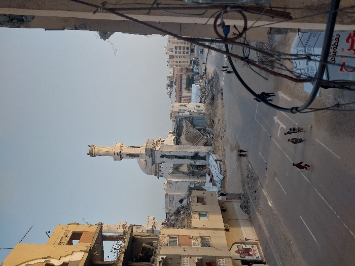 A cityscape devastated by war features a mosque with a towering minaret. Buildings are damaged or destroyed, rubble strewn about. A road runs through the scene with a few pedestrians walking along it. Power lines clutter the top of the frame, adding to the impression of destruction and urban decay. The sky is clear and pale.