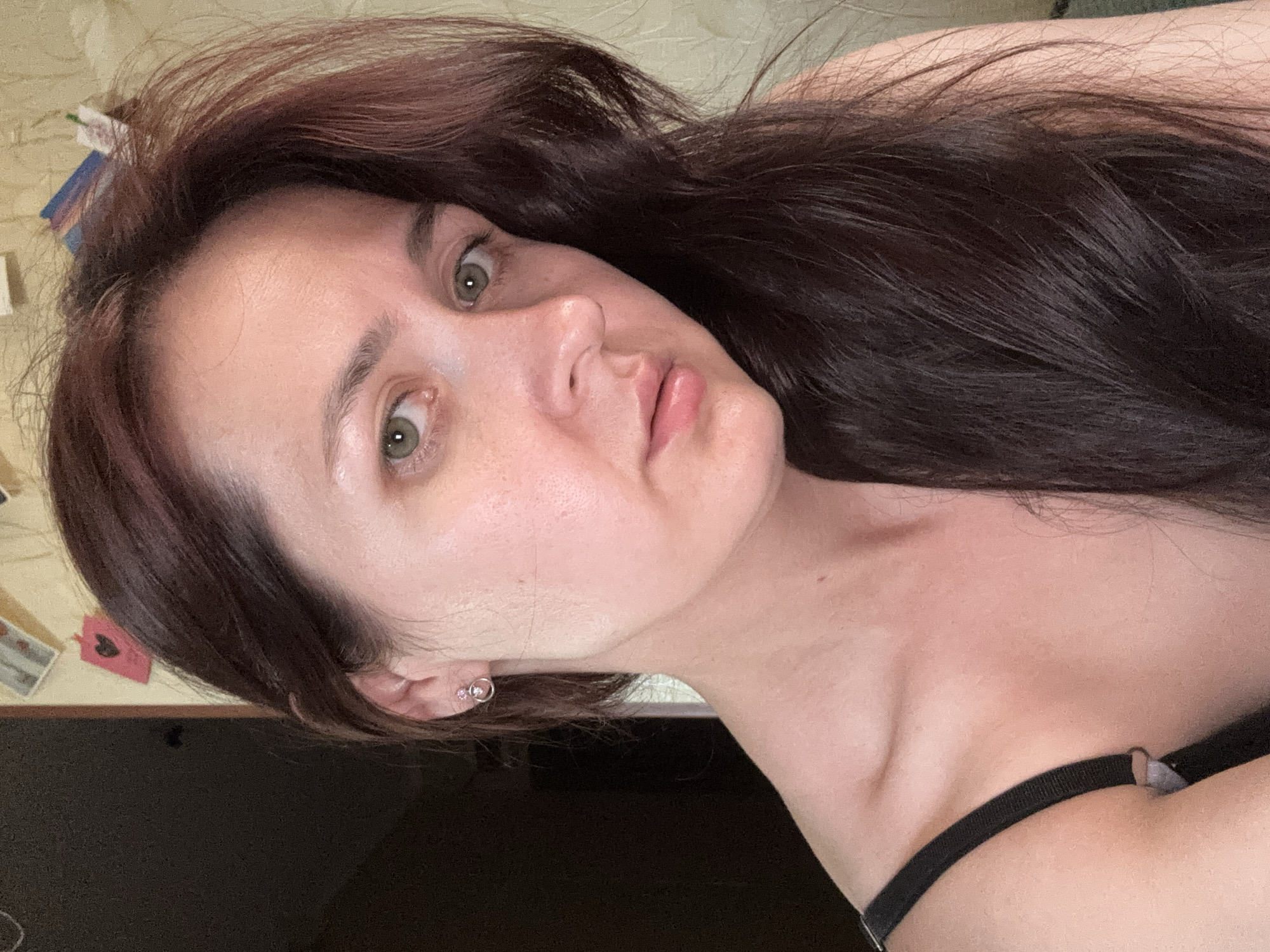 A close-up, head-and-shoulders portrait of a woman with long, dark auburn hair. She has fair skin, green eyes, and is wearing a black strap, possibly from a camisole or bra. A small, silver hoop earring with a pink gem is visible in her left ear. The background is out of focus, showing a wall with some decorative elements and a dark, out-of-focus area on the left. The lighting suggests an indoor setting, possibly a room in an apartment in Naberezhnye Chelny, Russia.