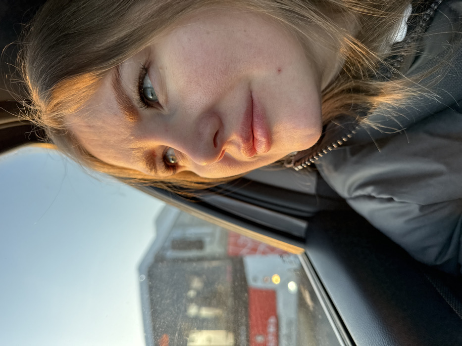 A young adult female is depicted in profile within the interior of a vehicle, looking rightward out a side window. Her light brown hair and the right side of her face are illuminated by warm, golden sunlight. Her eyes, appearing blue or green, are directed upwards and outwards, suggesting observation of the external environment. She exhibits a neutral to slightly contemplative expression. Her skin shows fine pores and a small, reddish mark on her lower right cheek. She is wearing a dark grey, possibly puffer-style, jacket with a zippered front and a visible white layer underneath. The vehicle's dark plastic door panel and window frame are visible in the foreground. Outside the window, a blurred white and red bus, consistent with public transport, is visible under a clear, pale blue sky. The bus has black-tinted windows and red horizontal stripes. The scene is located in the city of Froly, Russia.