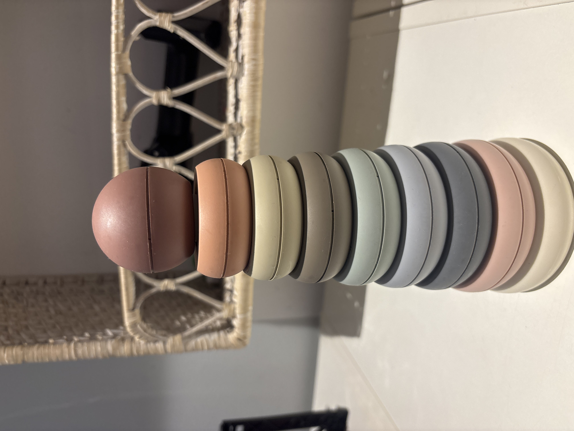 A stack of colorful silicone stacking rings is centered on a white surface, casting a shadow to the right. The rings are arranged from largest at the bottom to smallest at the top, with a spherical topper. The colors of the rings, from bottom to top, are: cream, blush pink, dark grey, light blue-grey, sage green, taupe, cream, and dusty rose. In the background, a woven shelf with decorative heart-shaped cutouts hangs on a light grey wall. The scene is brightly lit, suggesting an indoor setting. There are no people present in the image. The location is not identifiable as Dubai, United Arab Emirates.