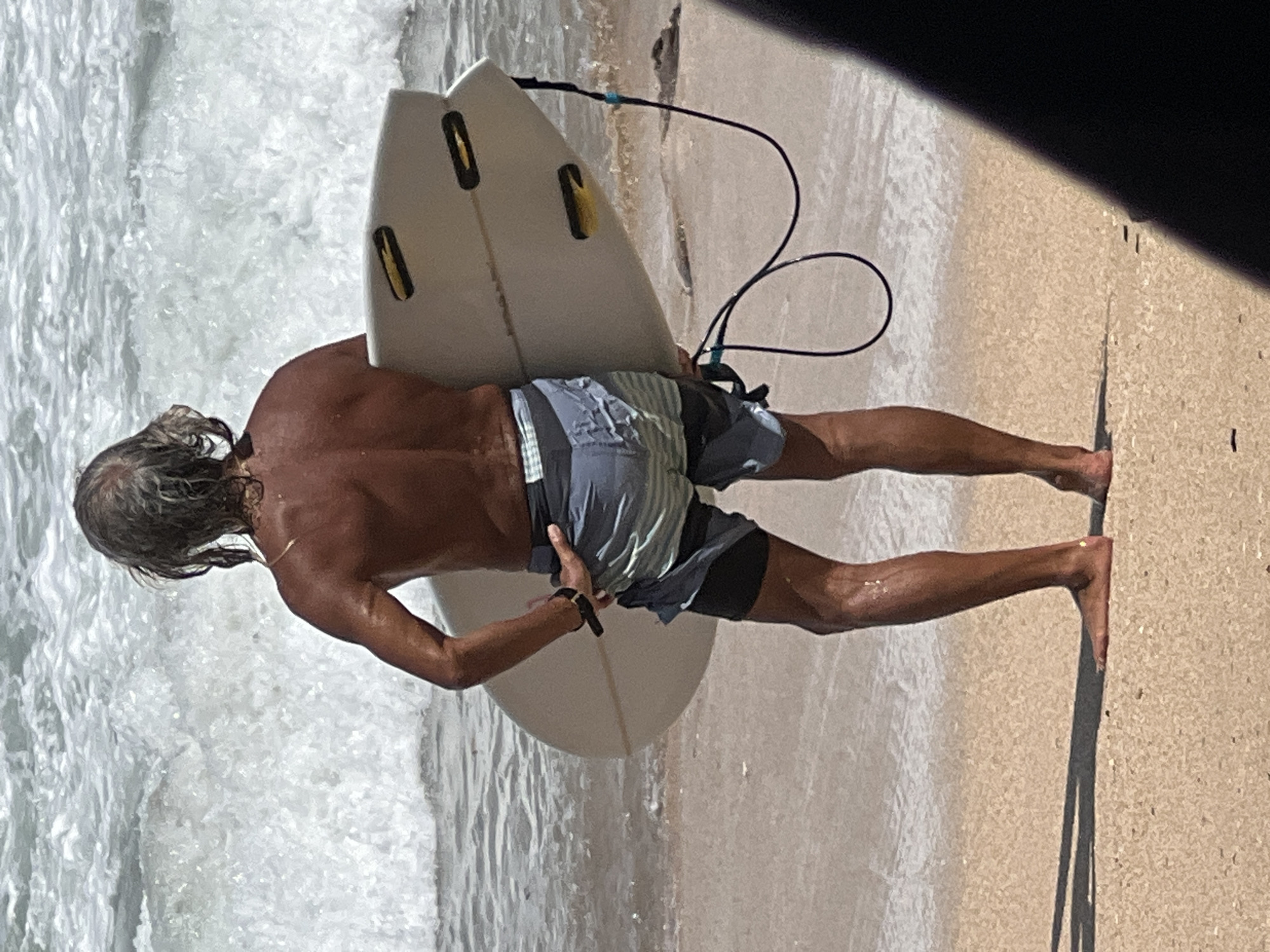 An adult male with long, graying hair stands on a sandy beach in Kuta, Indonesia, facing away from the camera and towards the ocean. He is holding a light-colored surfboard under his right arm. The surfer is shirtless, revealing a tanned physique, and is wearing patterned swim trunks over black shorts. The ocean behind him is choppy with white-capped waves. The ground is a light-colored sand, and the water's edge is visible with small waves lapping at the shore. A dark, blurred object occupies the lower right corner of the frame, obscuring part of the scene.