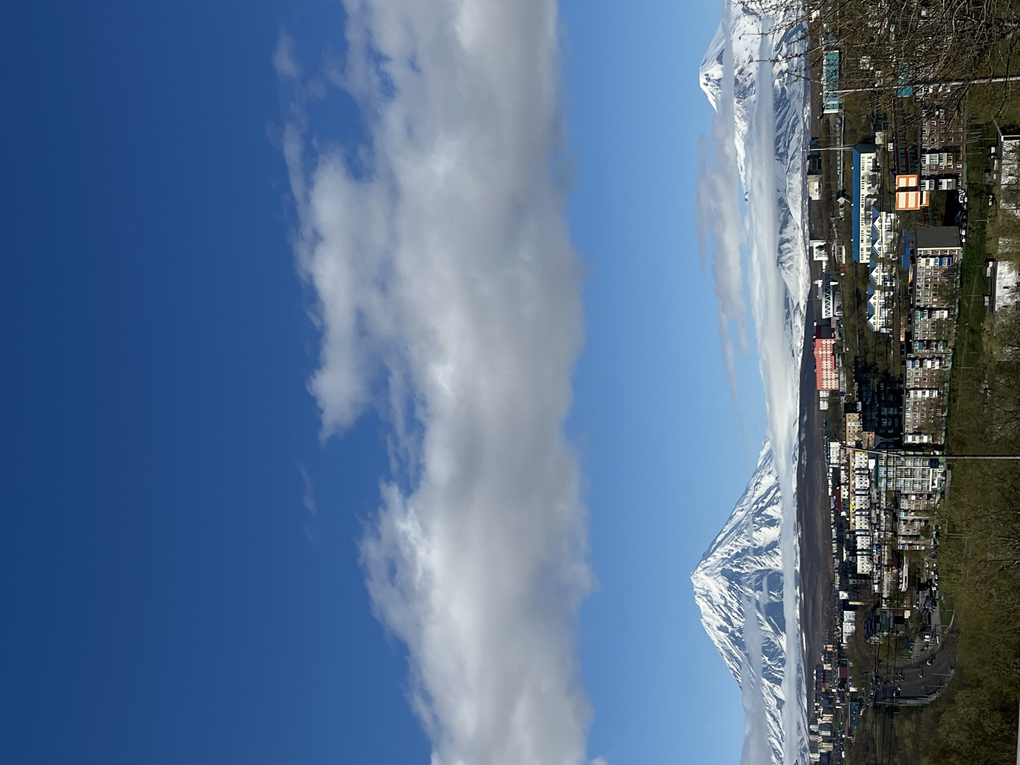 The image shows a view in Petropavlovsk-Kamchatsky, Russia. A large, snow-covered mountain dominates the right side of the frame, partially obscured by a vertical line of clouds. Below the mountain is a densely built-up area with residential buildings. A road winds along the base of the mountain. The left side of the frame is filled with a blue sky, with more clouds partially obscuring it.