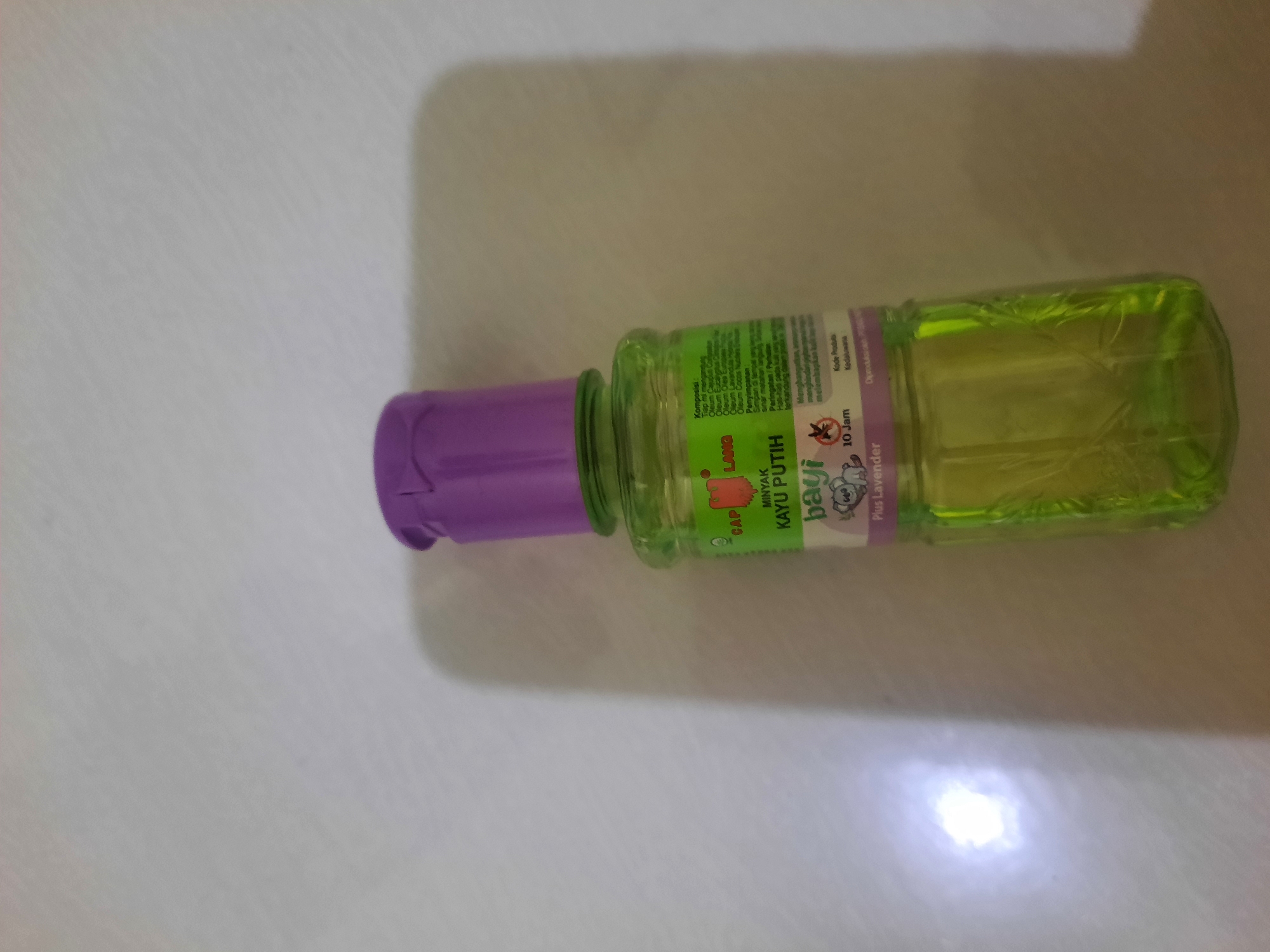 A product bottle with a green liquid and a purple cap stands upright on a light-colored, textured surface. The bottle has a label with text and an image of a koala. The label is partially obscured by the bottle's curvature and the lighting. The surface appears to be a wall or a table. A bright light source casts a shadow to the right of the bottle. There are no people or animals visible in the image. The setting is indoors. The product is "Minyak Kayu Putih Bayi Plus Lavender" (Baby White Wood Oil Plus Lavender).