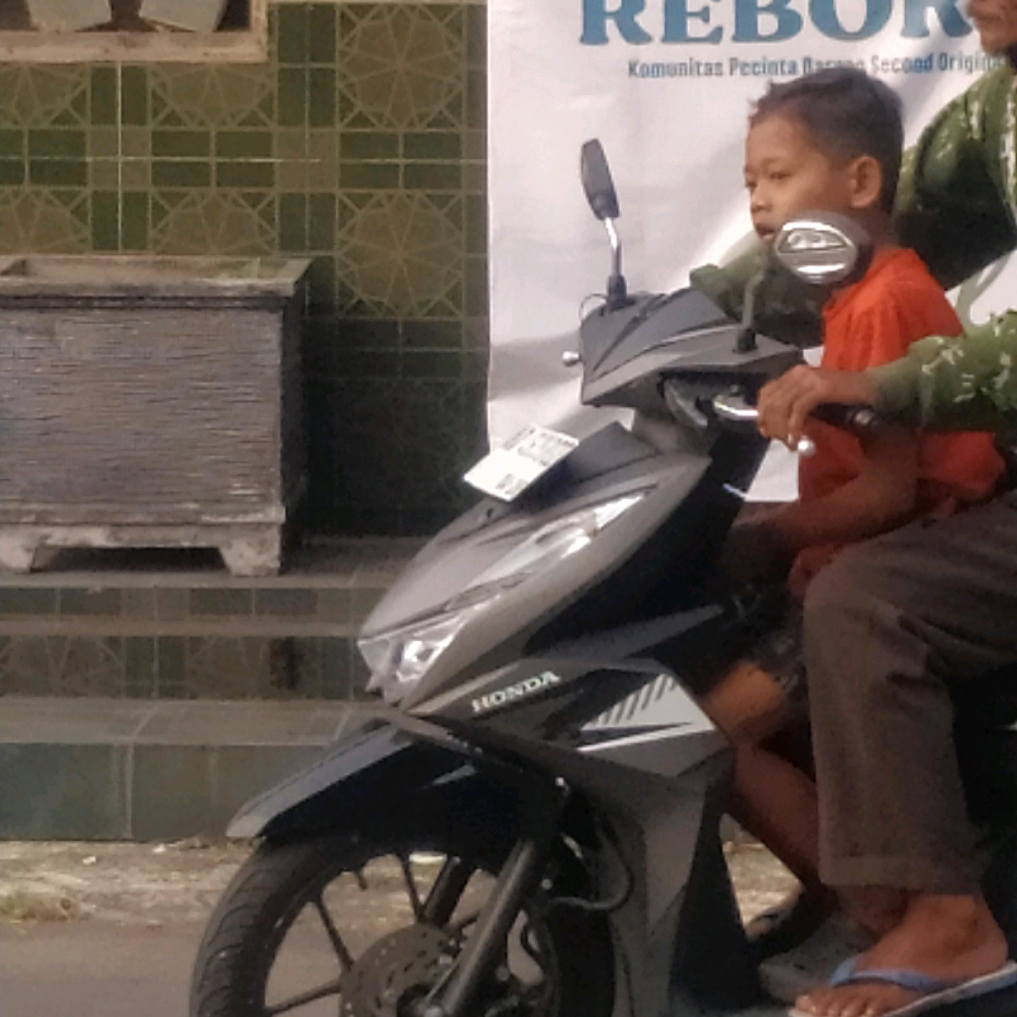 In Batan, Indonesia, a young boy wearing an orange shirt is seated on a gray motorcycle as a passenger. Behind him, an adult, partially visible, is also on the motorcycle, wearing a green patterned garment and brown pants. The adult's hands are on the handlebars. The boy is looking to his left. The motorcycle has a "HONDA" logo on its front panel. In the background, a building with green tiled walls and a white banner with blue text is visible. Part of a large, gray, rectangular object, possibly a planter or a bench, is in the foreground to the left. The ground is paved, and there is some debris scattered on it.