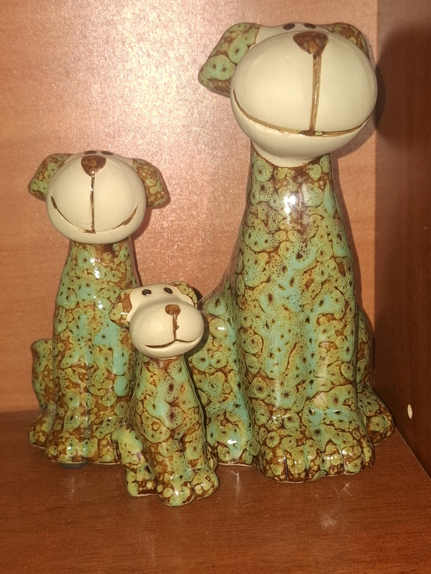 Three ceramic dog figurines are arranged on a wooden shelf. The largest figurine stands in the back right, with two smaller figurines positioned in front of it, one slightly to the left and another to the right. All figurines share a similar design, featuring light-colored heads with brown accents for noses, eyes, and mouths, and bodies adorned with a speckled green and brown glaze. The background is a textured, light-pink surface. The shelf itself is made of wood with a visible grain. No specific location in Almaty, Kazakhstan is discernible from the image.