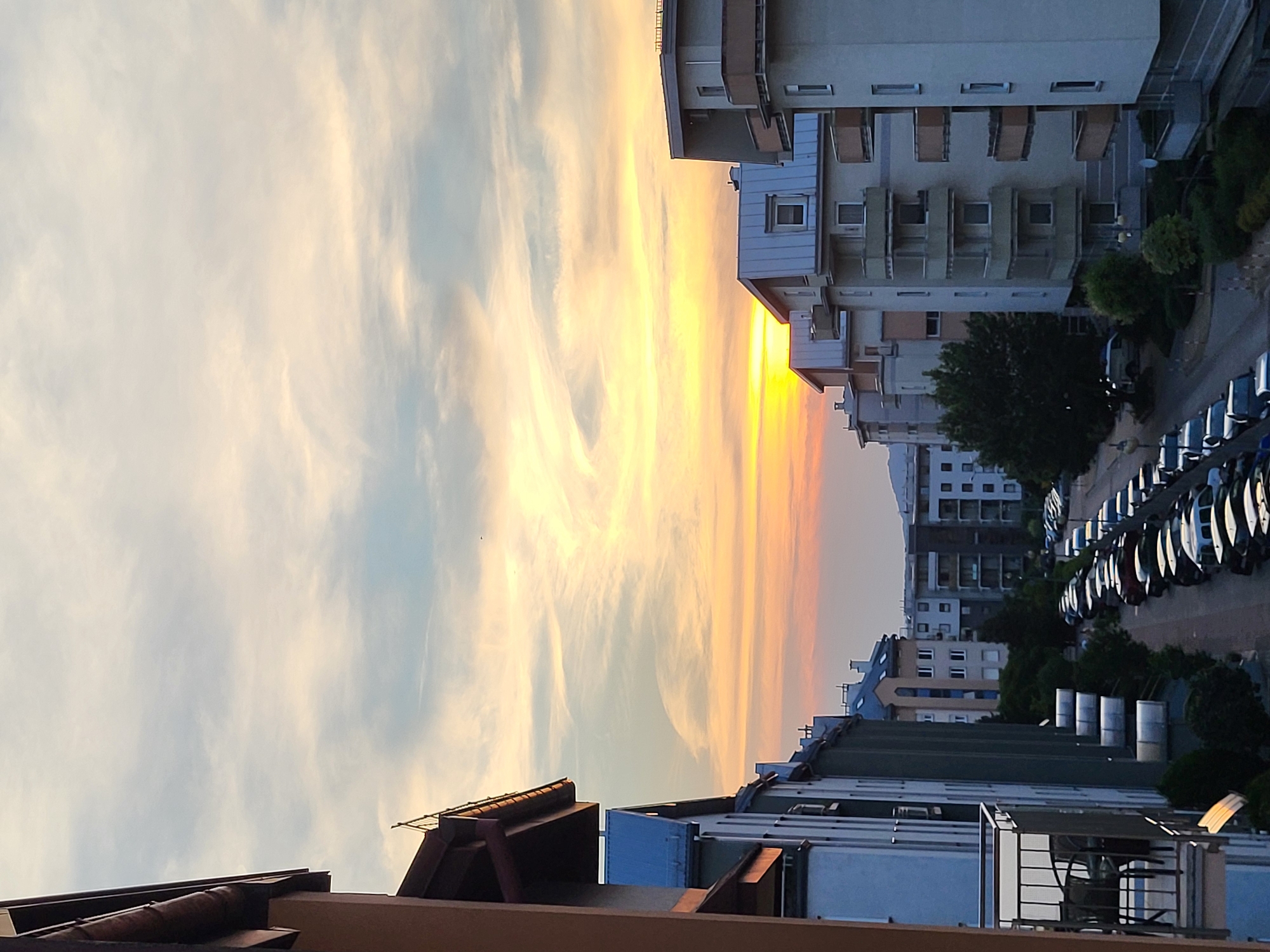 The image depicts a street view in a city during sunset. The sky is a vibrant gradient of orange and yellow, with wispy clouds. On the left side of the frame, a building with a brown roofline and balconies is visible, with other residential buildings extending into the background. The buildings are a mix of white, light blue, and gray, with multiple stories and balconies. A street lined with parked cars runs down the center of the image, with trees and other greenery on either side. The overall scene conveys a sense of urban tranquility as the day ends.