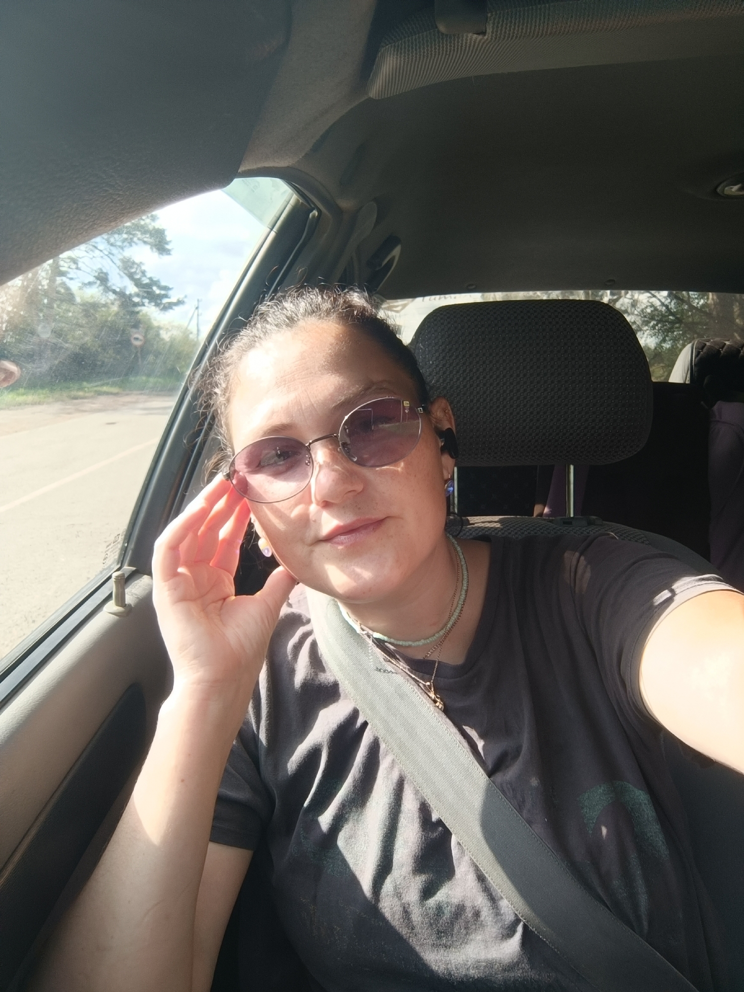 A middle-aged woman is captured in a selfie taken from inside a car, which is traveling on a road in or near Foki, Russia. The woman, wearing sunglasses and a dark t-shirt with a necklace, has her hand resting on her cheek. The car's interior is visible, including the seatbelt, headrest, and a portion of the window. Outside the window, trees and a road are visible, suggesting a rural or suburban setting. The lighting indicates it is daytime, with sunlight streaming into the car.
